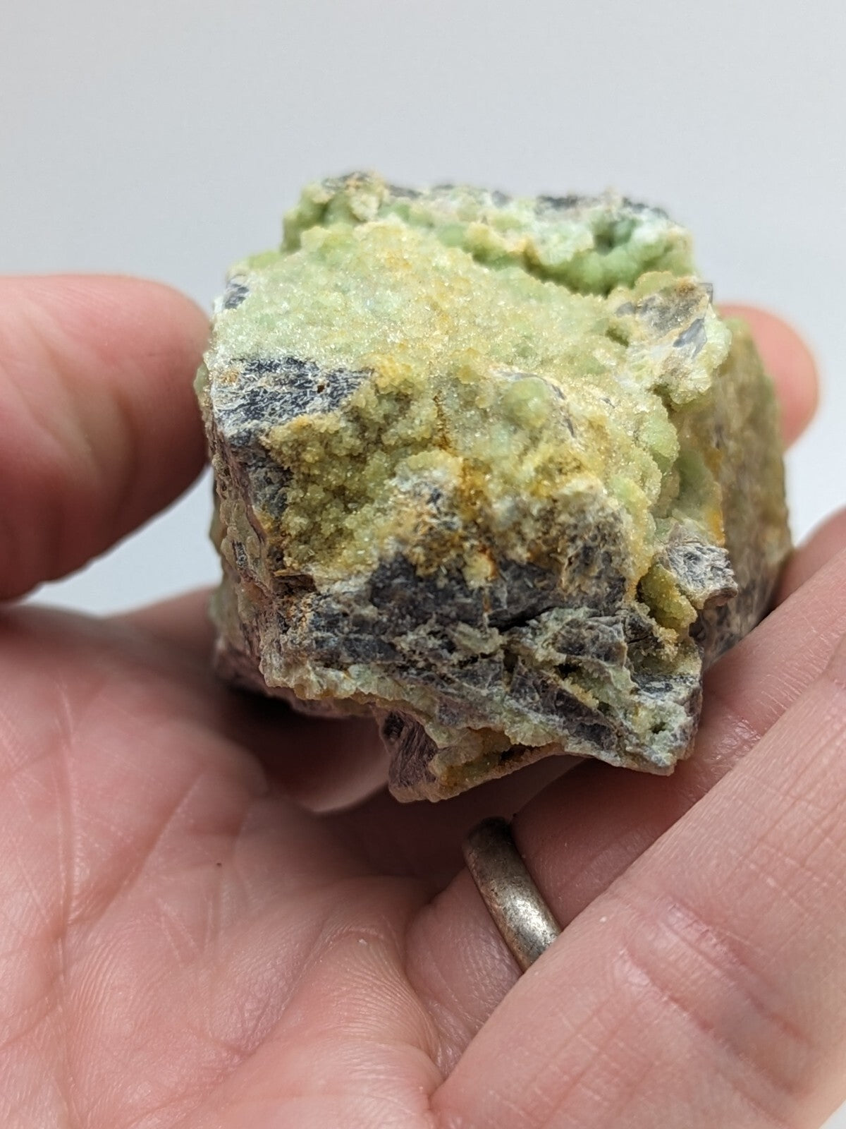 A rough, irregularly shaped mineral specimen with a textured surface displaying patches of pale green and yellowish hues against darker gray matrix.