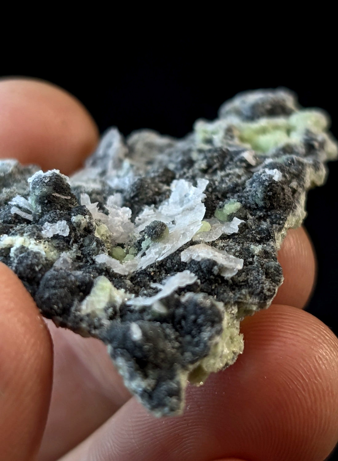 A hand holds a dark gray mineral specimen with translucent white and pale green crystalline formations.