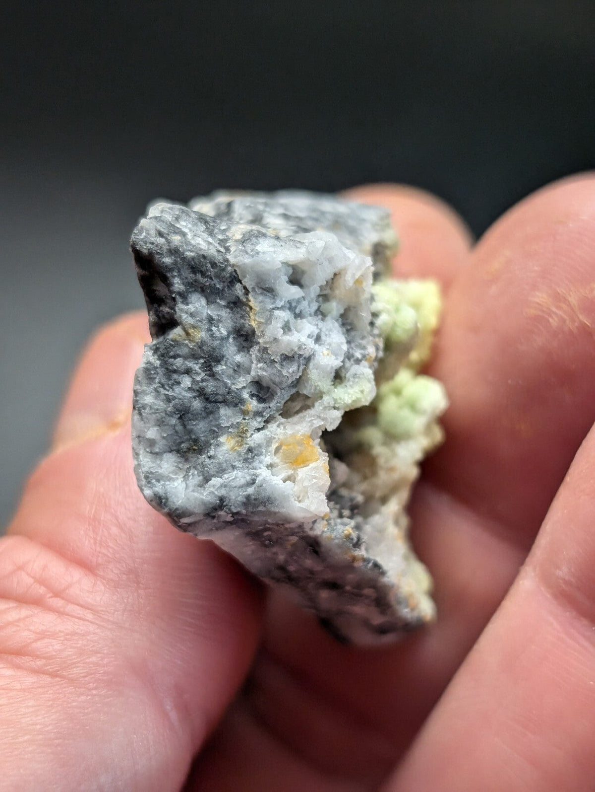 A hand holds a rough, irregularly shaped mineral specimen featuring gray and white crystalline textures with patches of pale green and yellowish hues.