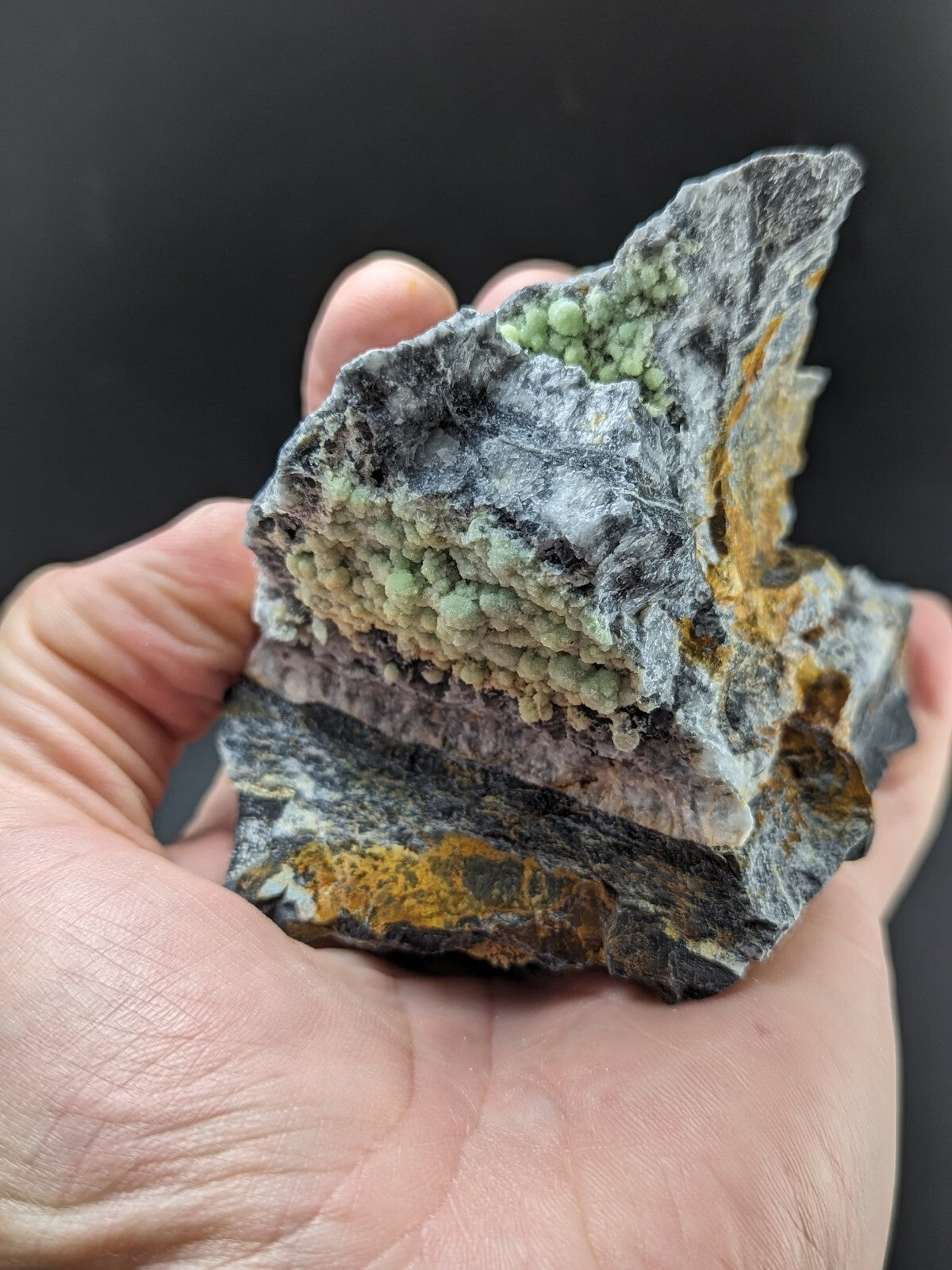 A hand holds a rugged, dark gray rock specimen featuring vibrant green crystalline formations and streaks of yellow mineral.