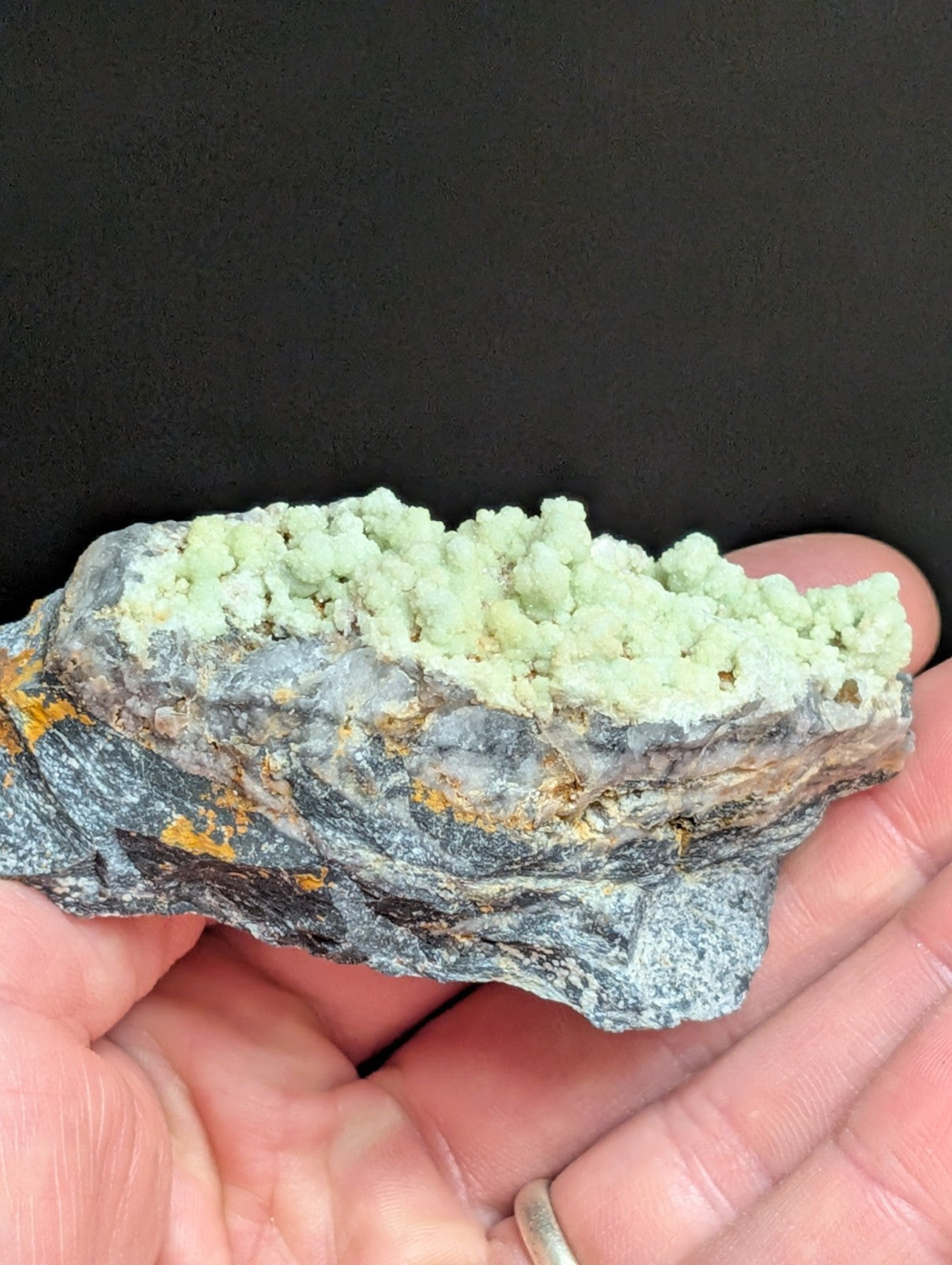 A hand holds a geological specimen featuring a grayish rock base with patches of orange mineral and topped by a cluster of pale green, crystalline formations.