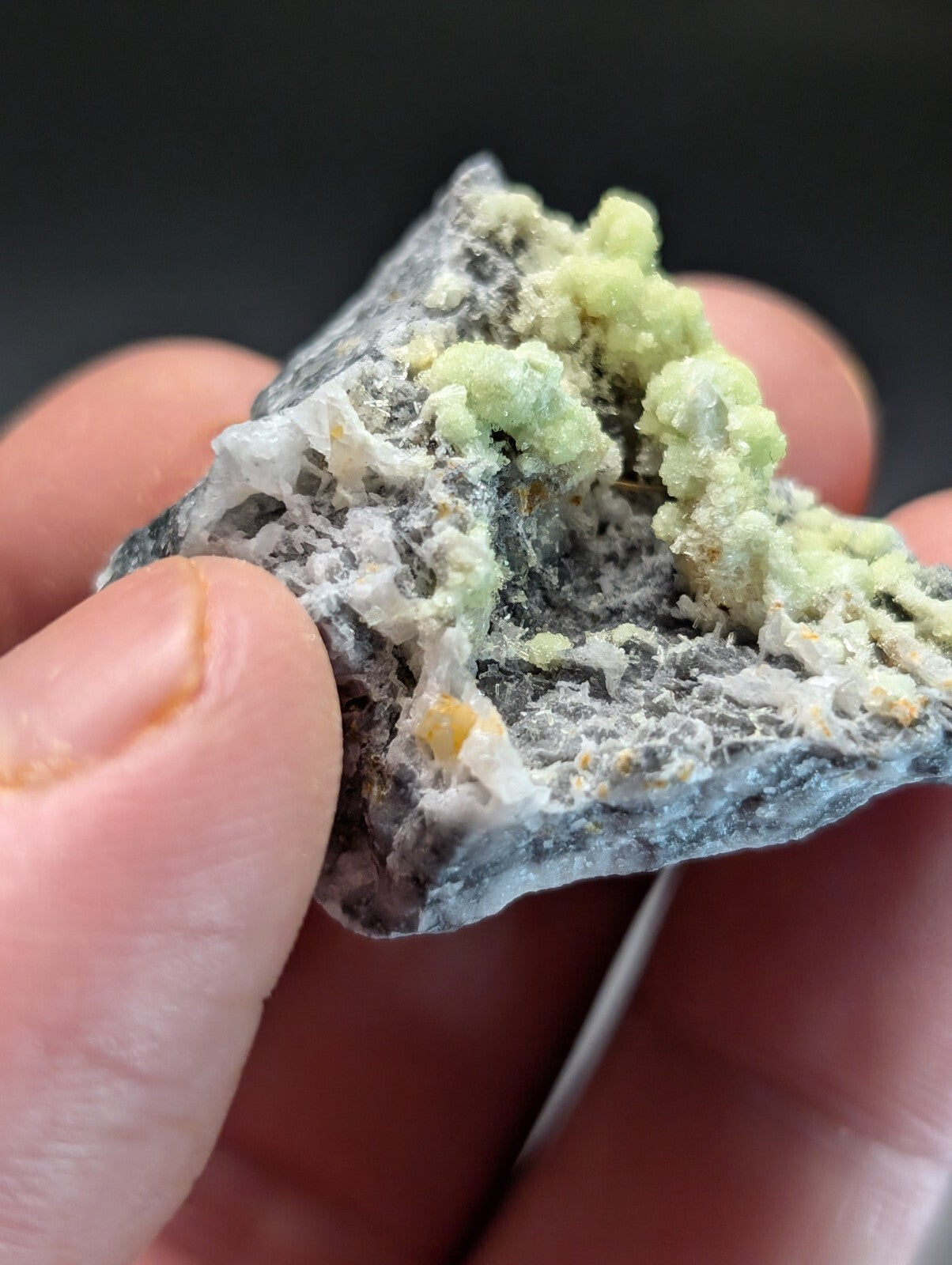 A hand holds a rough, irregularly shaped mineral specimen featuring clusters of pale green crystals growing from a dark gray and white matrix.