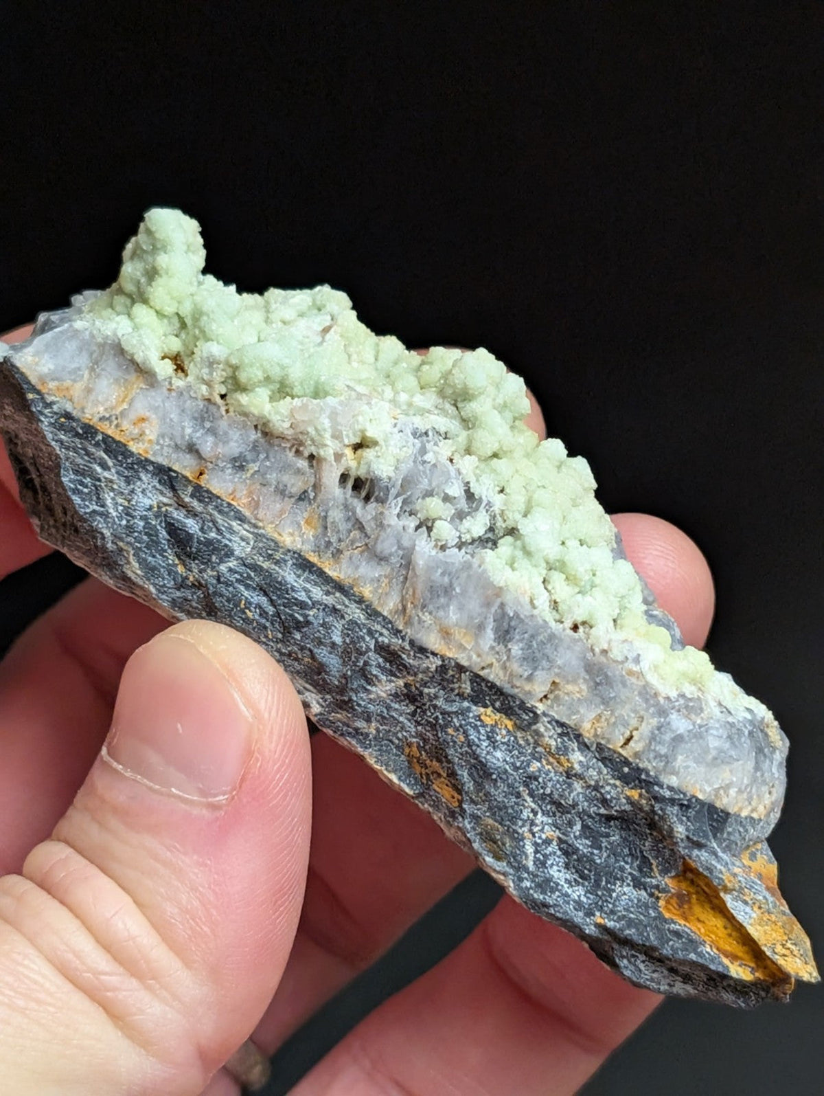 A hand holds a geological specimen featuring a dark gray, textured rock base with translucent quartz crystals and vibrant light green mineral formations on top.