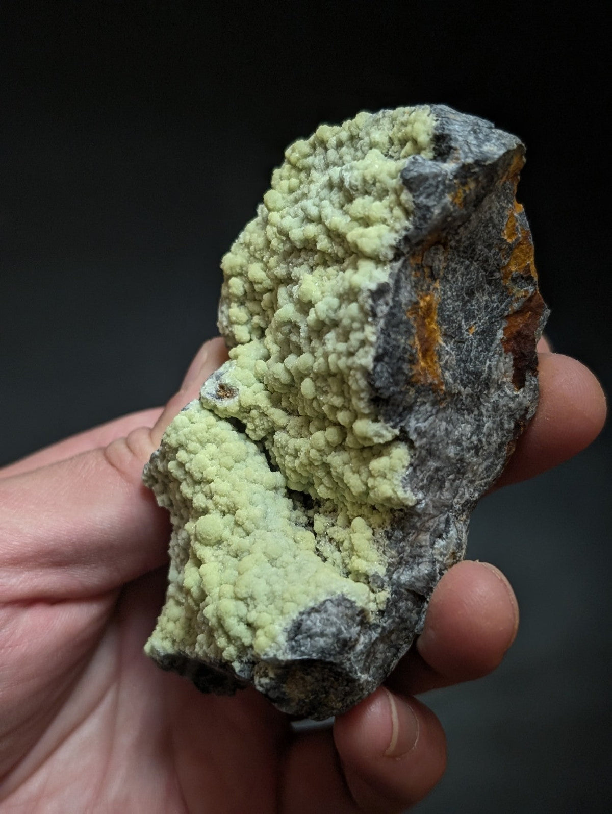 A hand holds a rough, irregularly shaped mineral specimen featuring vibrant pale green crystalline formations growing over darker gray rock with patches of orange and reddish hues.