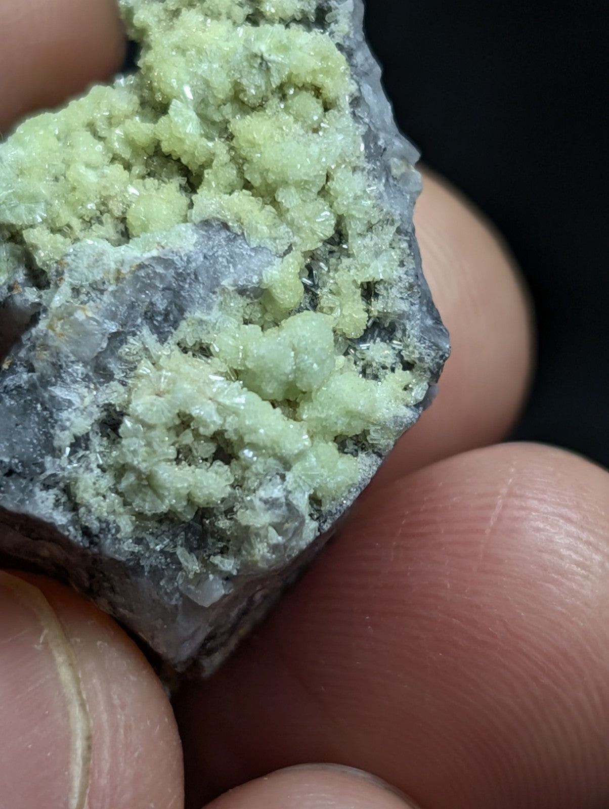 A hand holds a rough, irregularly shaped mineral specimen featuring clusters of pale green crystalline formations growing on a darker gray rock base.