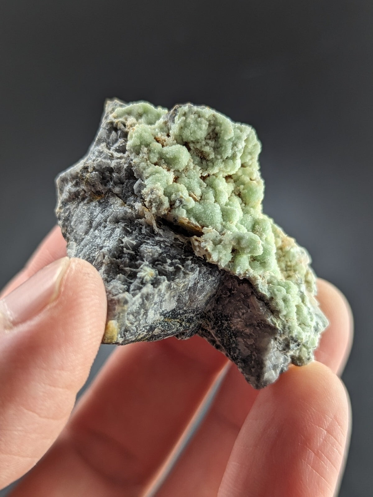 A hand holds a rough, irregularly shaped mineral specimen featuring a striking contrast between its dark gray base and vibrant light green crystalline formations.