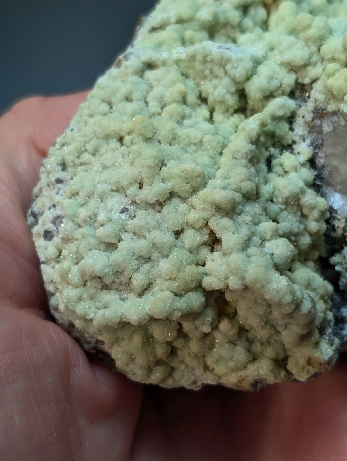 A hand holds a rough, irregularly shaped mineral specimen featuring dense clusters of pale green crystals with subtle hints of yellow and white.