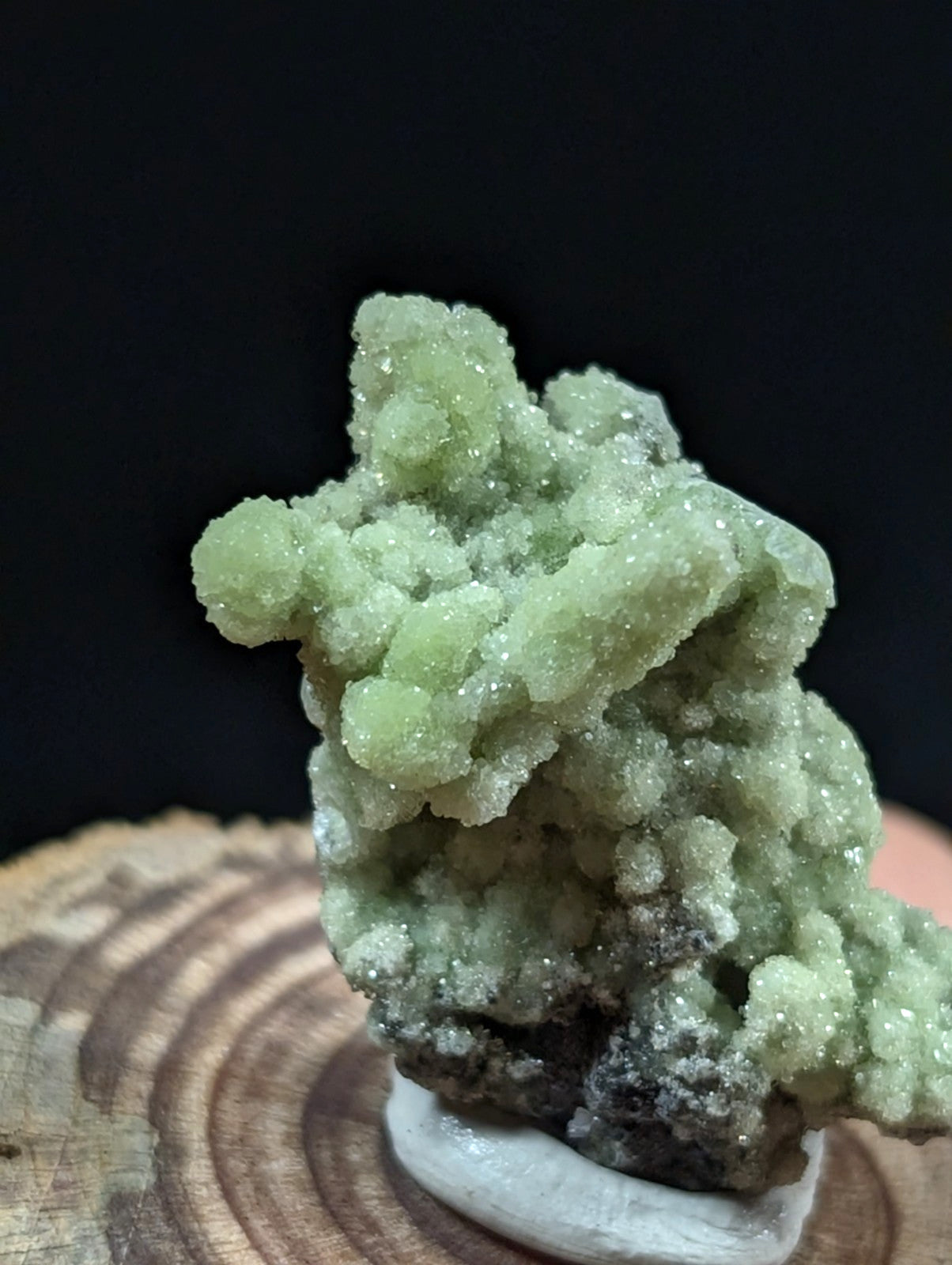 A pale green, crystalline mineral specimen with a textured, bumpy surface and some darker grayish base material.