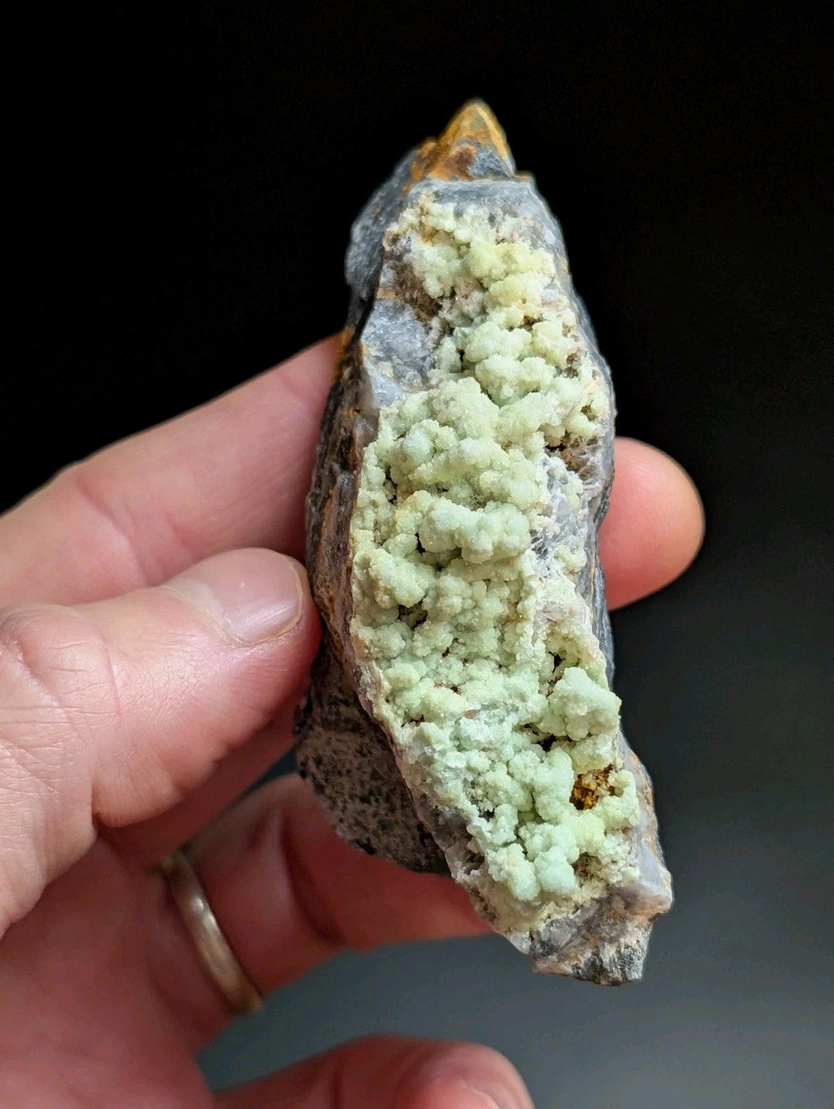 A hand holds a rough, irregularly shaped mineral specimen featuring a striking light green crystalline formation growing from a darker gray rock matrix.