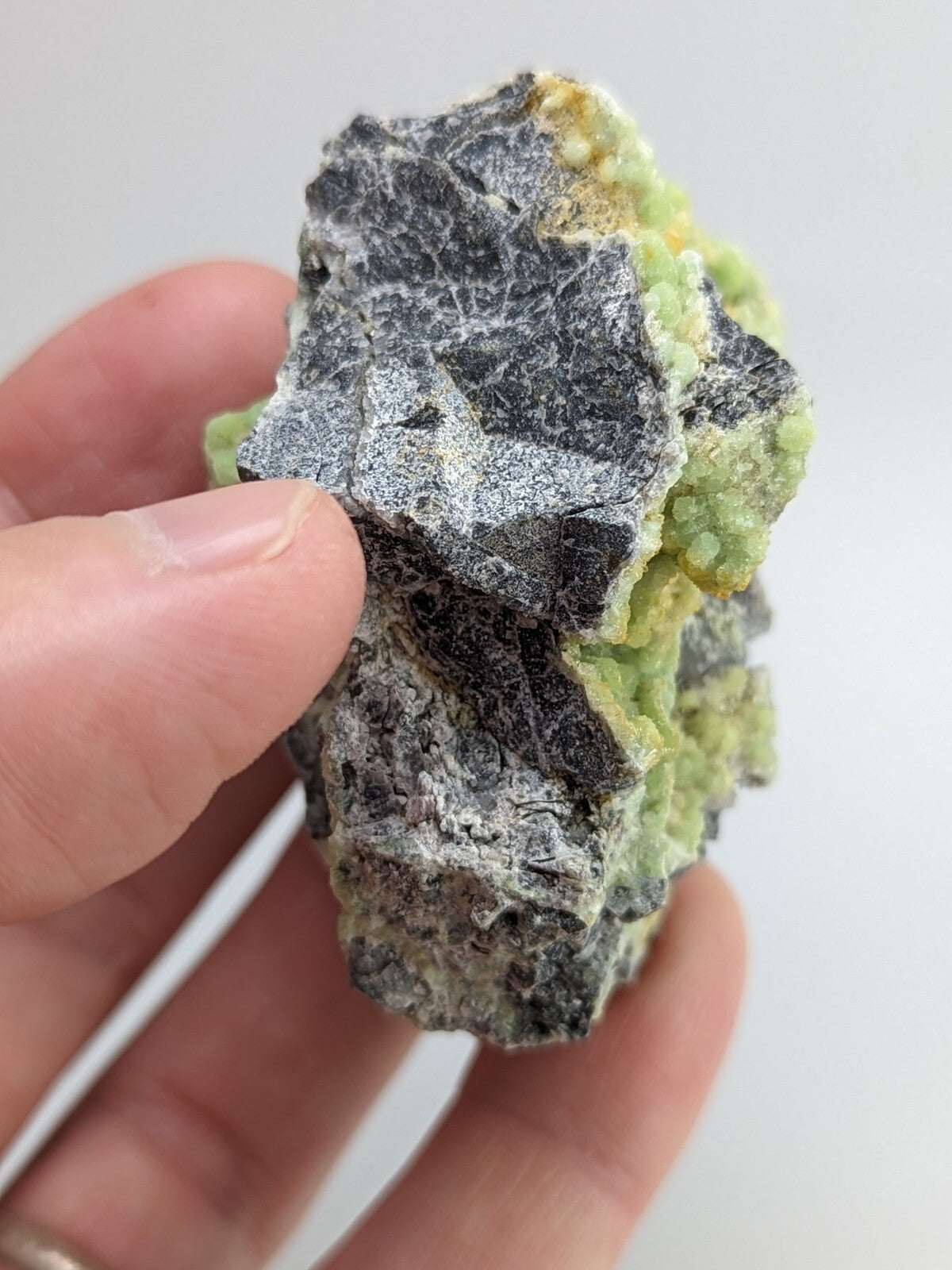 A hand holds a rough, irregularly shaped mineral specimen featuring dark gray and black crystalline sections contrasted with vibrant green patches.