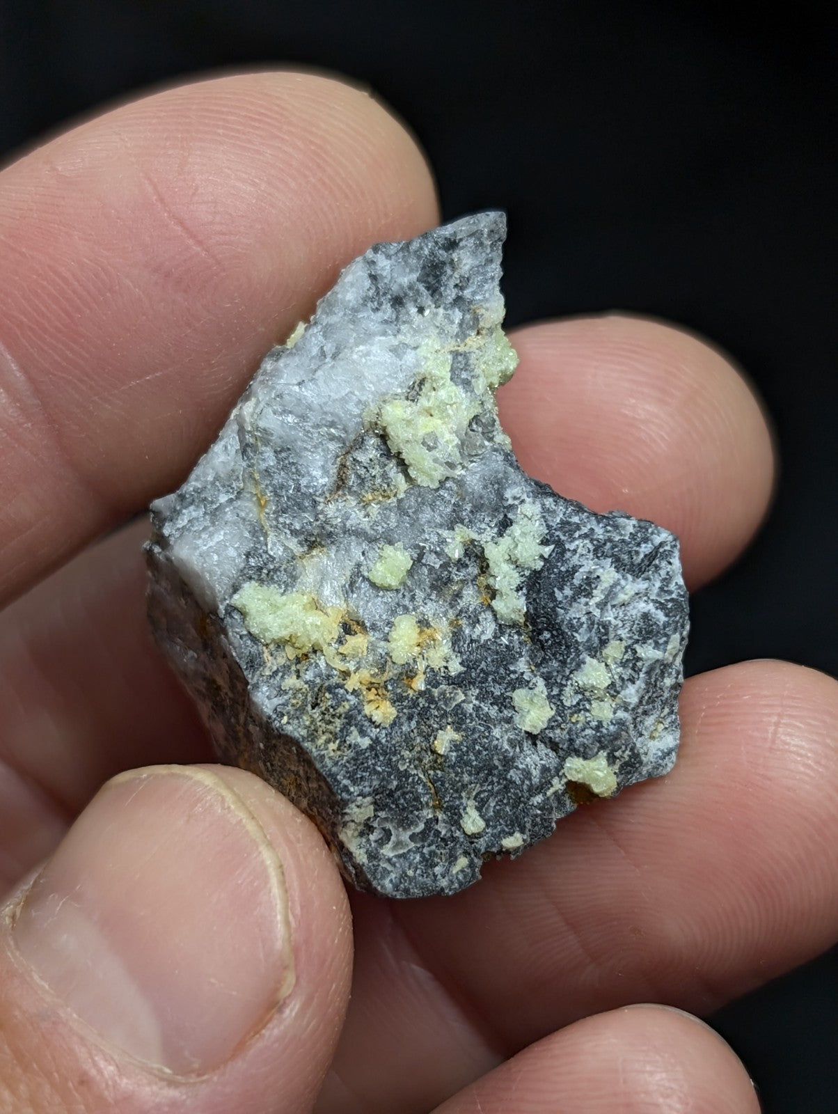 A rough, irregularly shaped mineral specimen with a dark gray base and patches of pale greenish-yellow crystalline formations.
