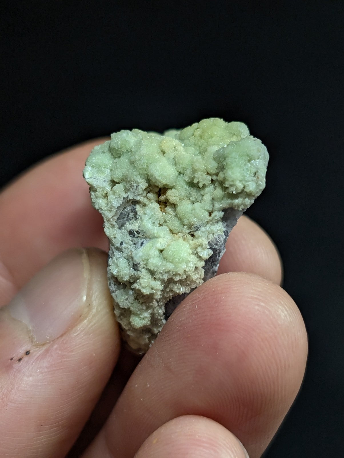 A textured, pale green mineral specimen with a rough, crystalline surface held between fingers.