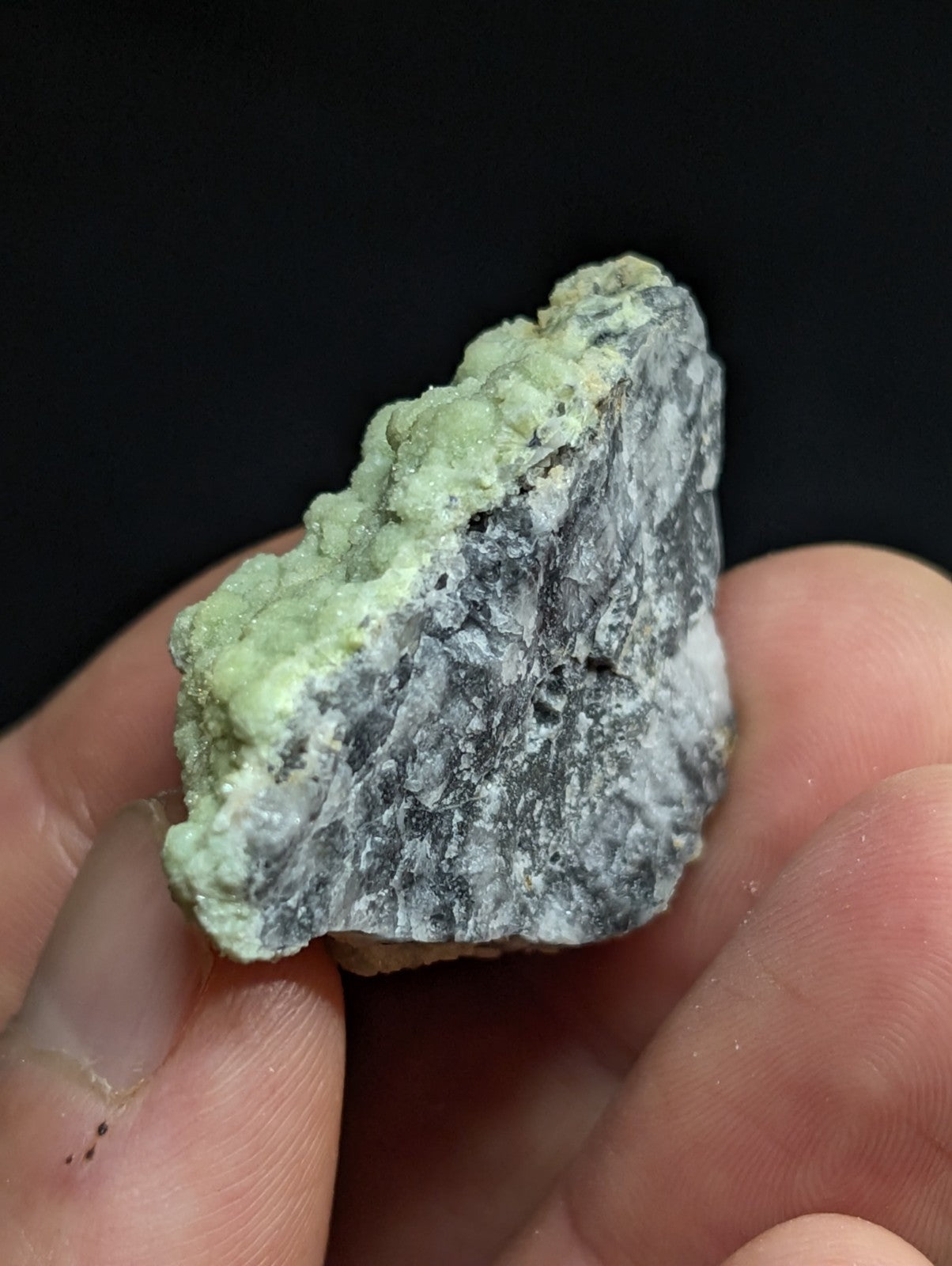 A hand holds a rough, irregularly shaped mineral specimen with a mottled surface of pale green and grayish-white hues.