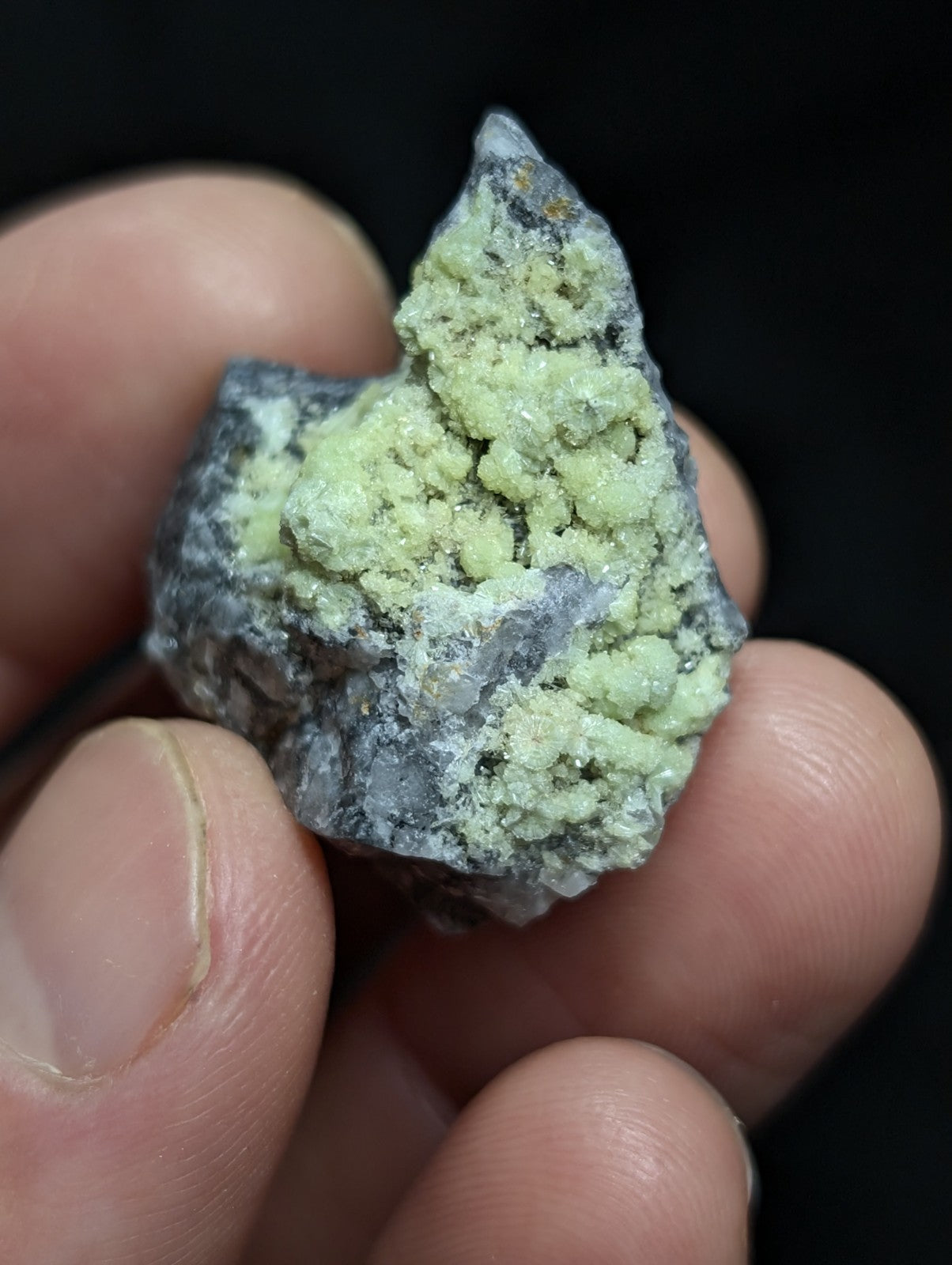 A hand holds a rough, irregularly shaped mineral specimen featuring patches of pale green crystalline growths over a darker grayish matrix.