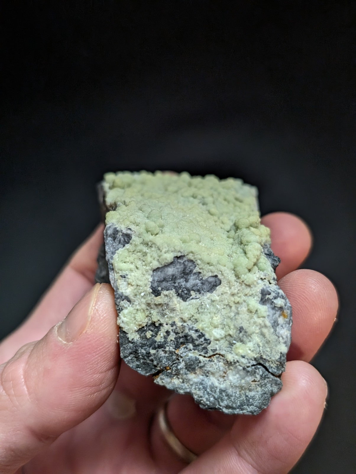 A hand holds a rough, irregularly shaped mineral specimen with a textured surface featuring patches of pale greenish-yellow crust over darker gray and black rock.