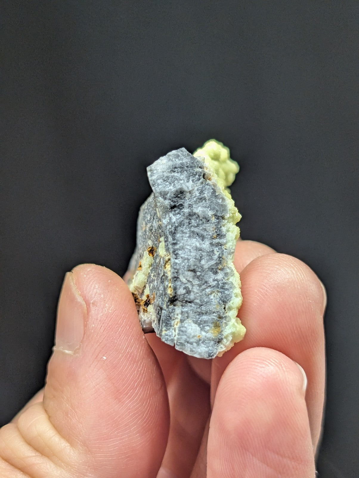 A hand holds a gray, rough-textured mineral specimen with patches of pale green crystalline growths.