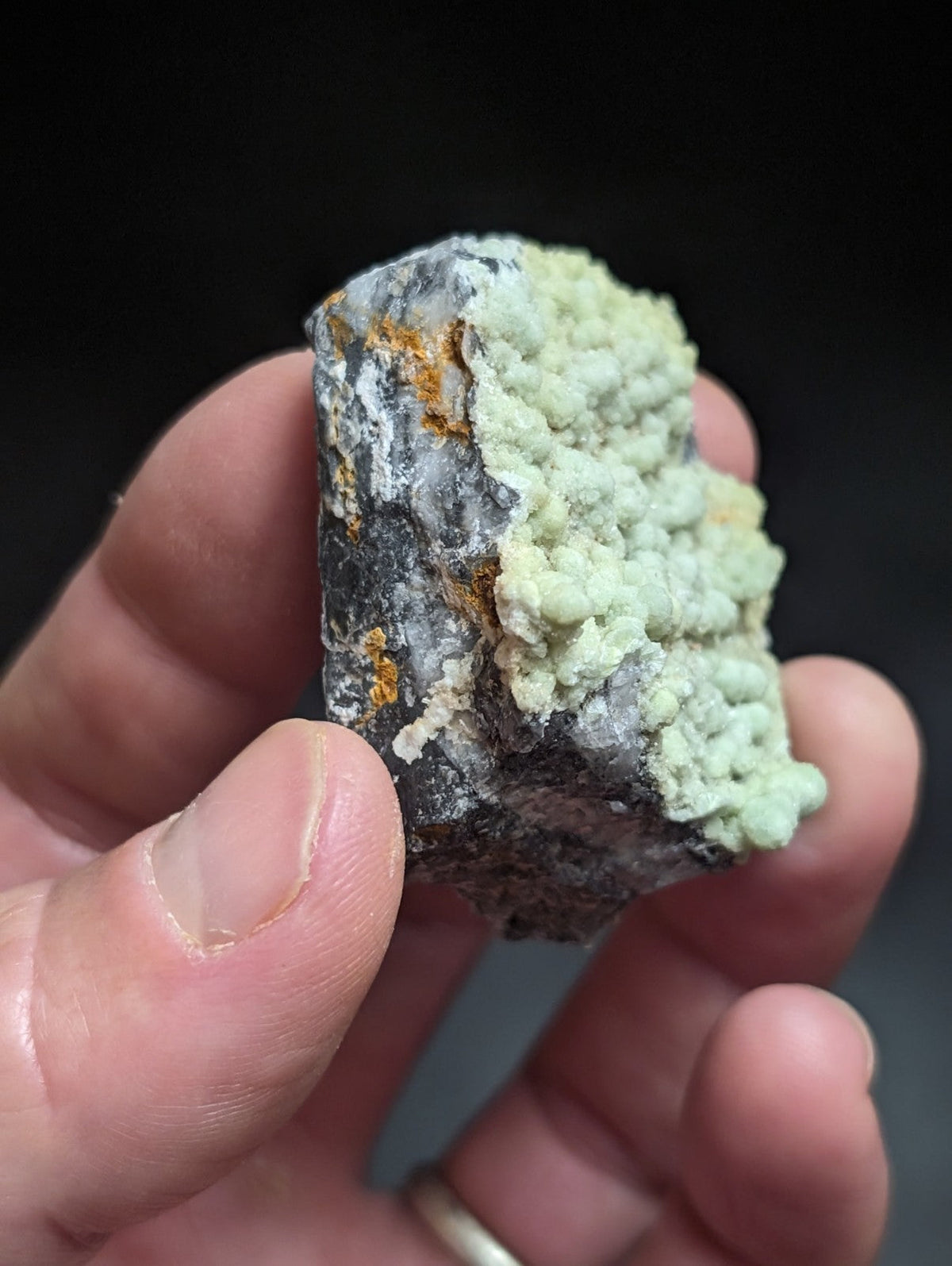 A hand holds a rough, irregularly shaped mineral specimen featuring a striking contrast between its dark gray base and vibrant light green crystalline formations.