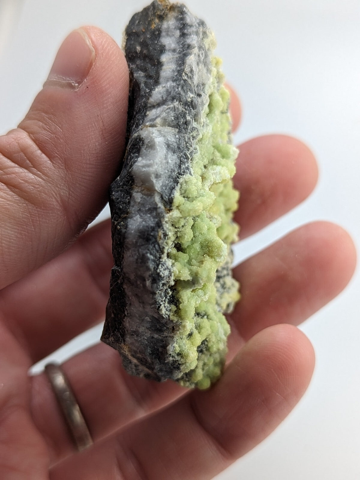 A hand holds a rough, irregularly shaped mineral specimen featuring vibrant green crystals growing from a dark gray matrix.