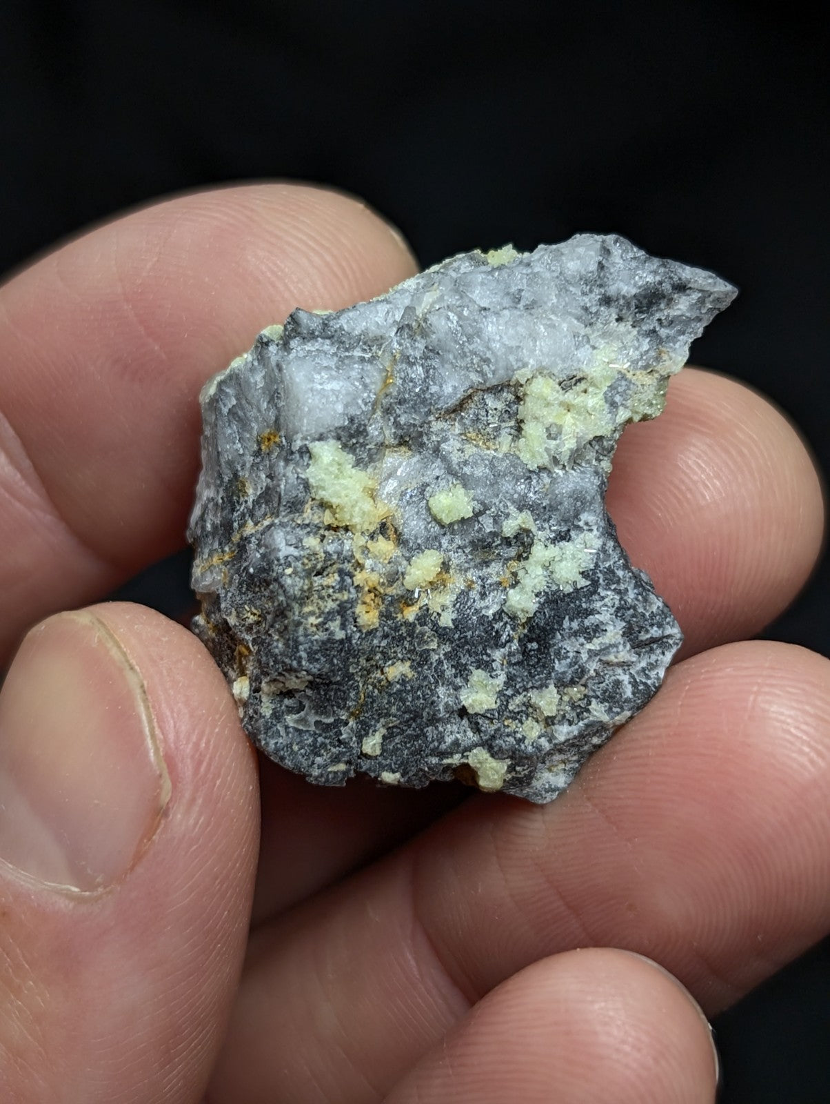 A rough, irregularly shaped mineral specimen with a dark gray base and patches of pale yellowish-green crystalline formations.