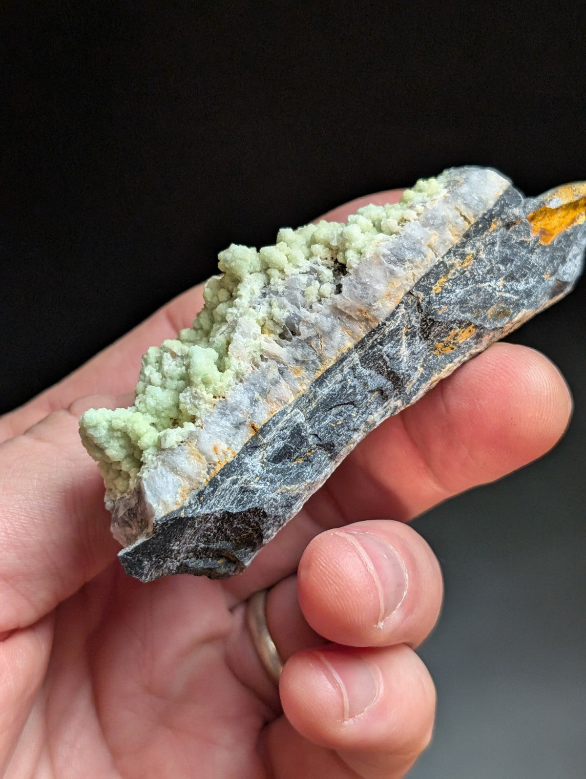 A hand holds a natural mineral specimen featuring a textured, light green crystalline formation atop a dark gray rock base with subtle orange streaks.