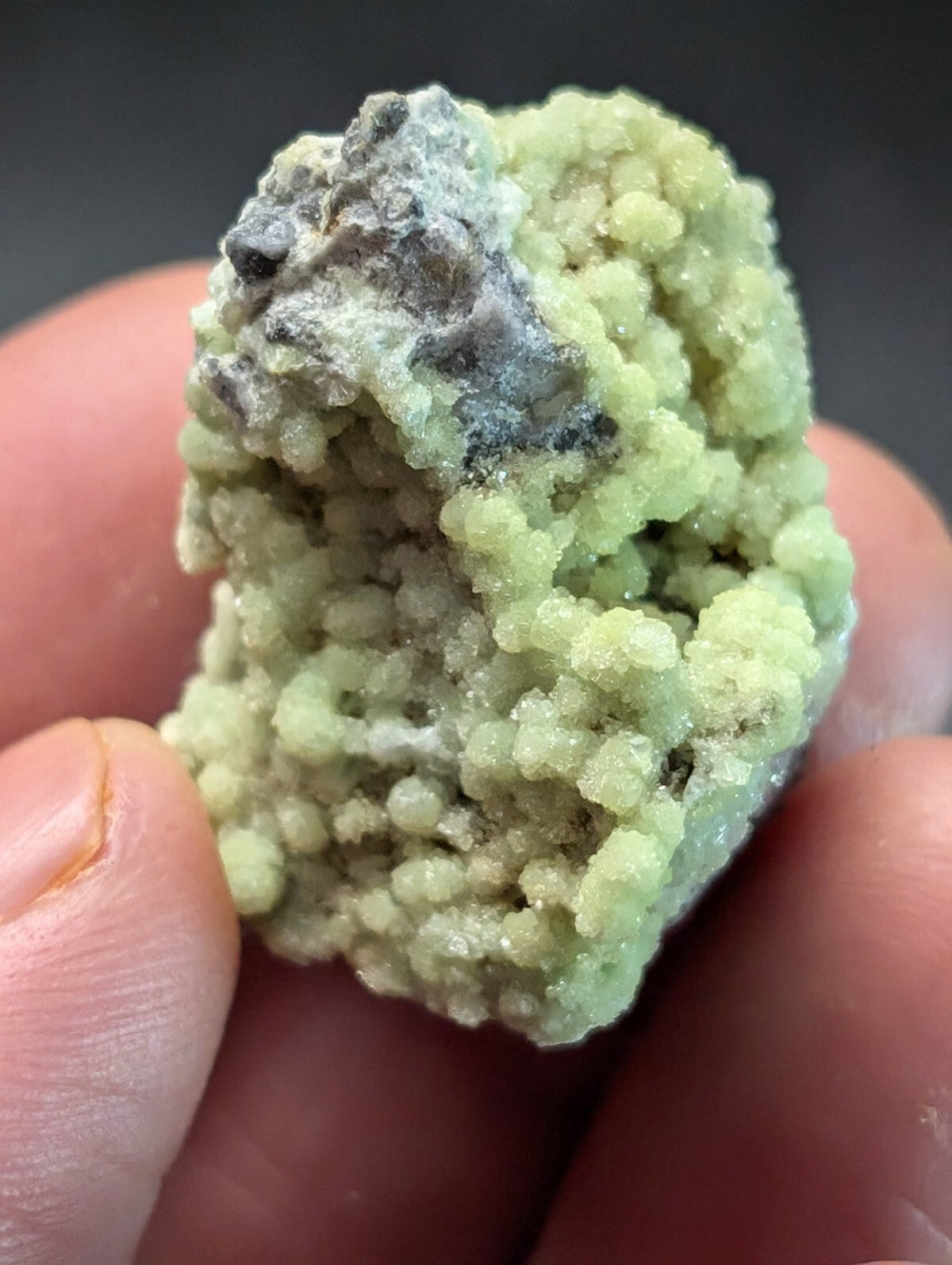 A pale green, crystalline mineral specimen with a textured surface and patches of grayish matrix.