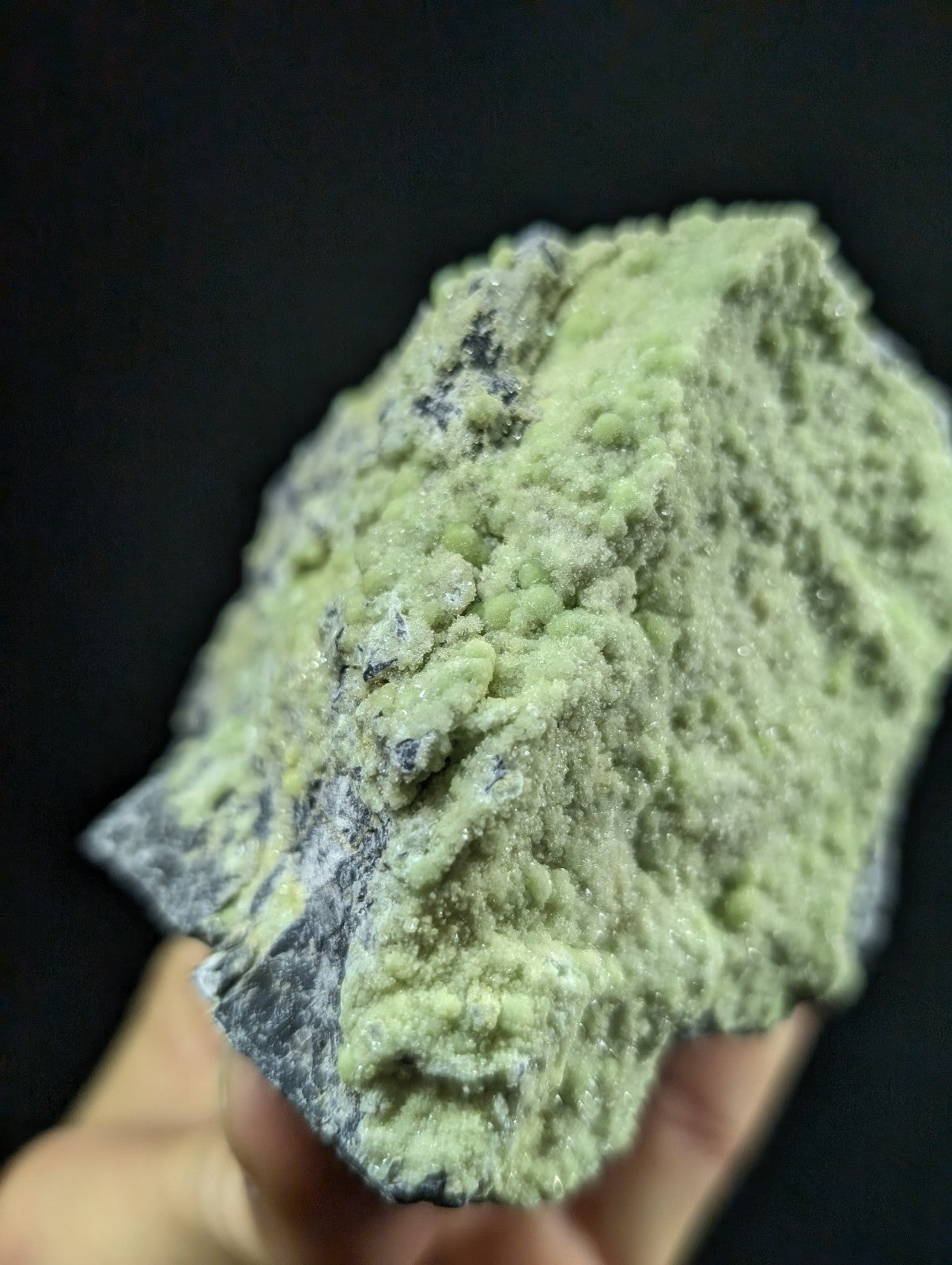 A rough, irregularly shaped mineral specimen with a pale green crystalline surface and darker grayish-black patches.