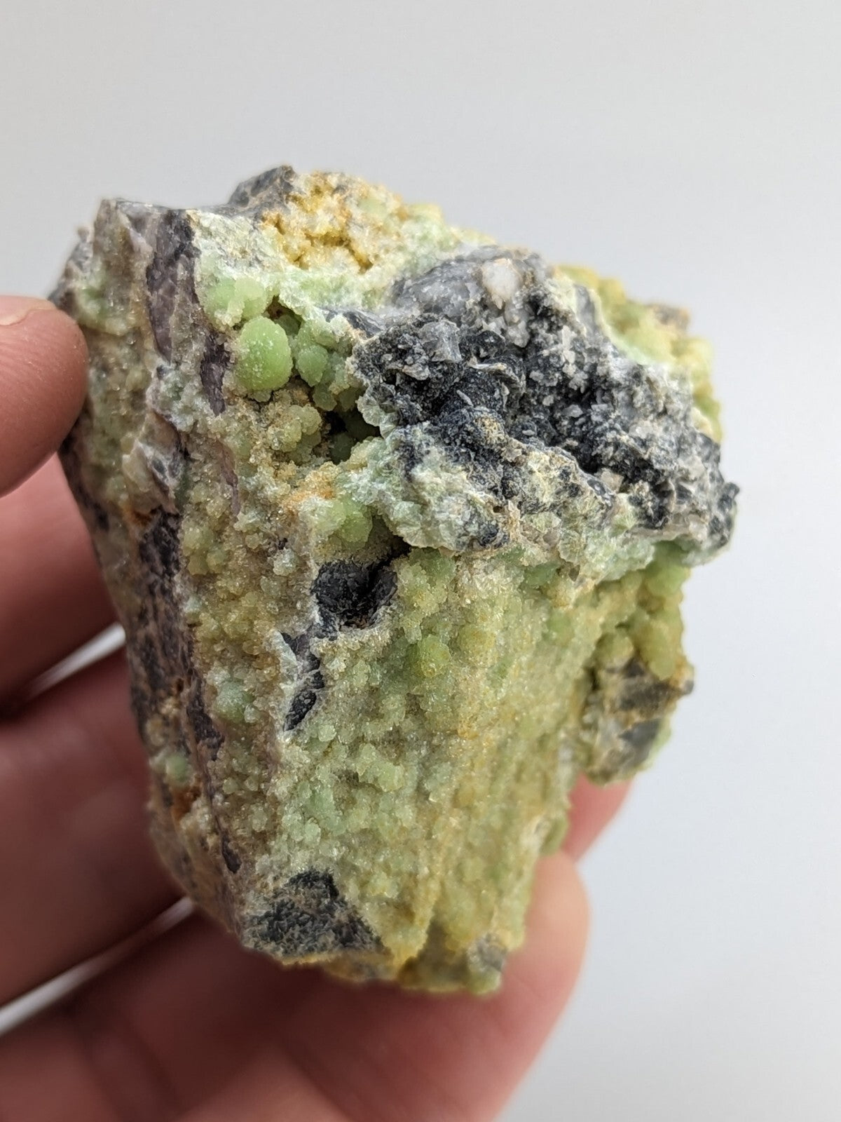 A hand holds a rough, irregularly shaped mineral specimen featuring clusters of translucent green crystals embedded in a matrix of dark gray and pale yellowish rock.