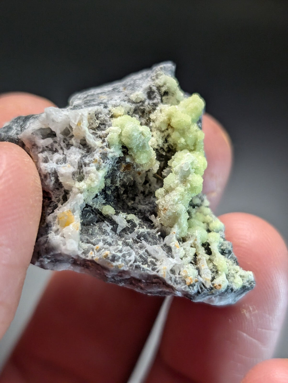 A hand holds a rough, irregularly shaped mineral specimen featuring vibrant green crystalline formations against a dark gray matrix with white veining.