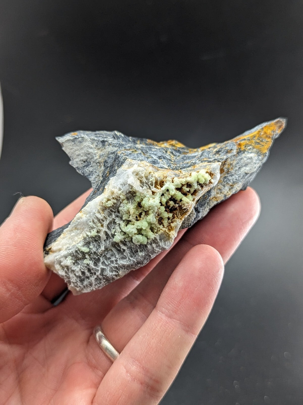A hand holds a rugged, irregularly shaped mineral specimen featuring a dark gray base with patches of vibrant yellow and delicate light green crystalline formations.