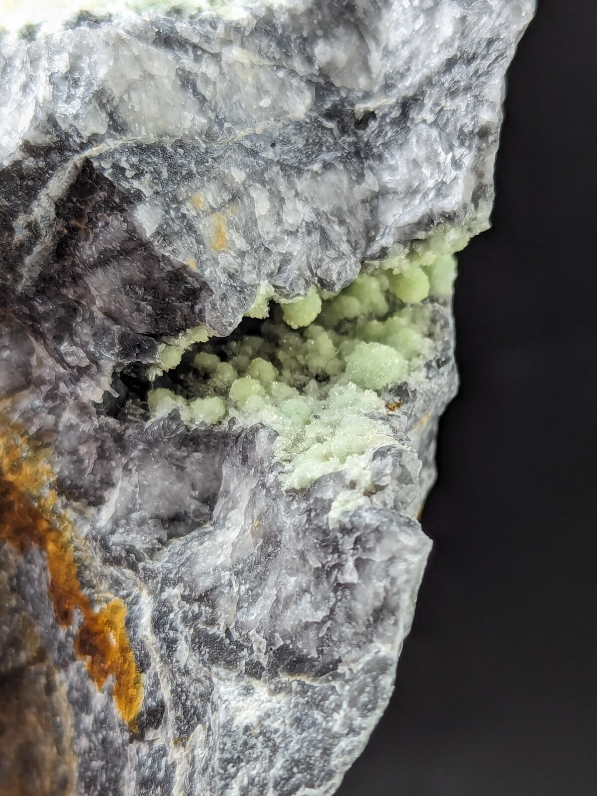 A striking mineral specimen featuring a gray, crystalline rock matrix with vibrant green, bead-like crystals growing from its interior and orange-yellow streaks along the surface.