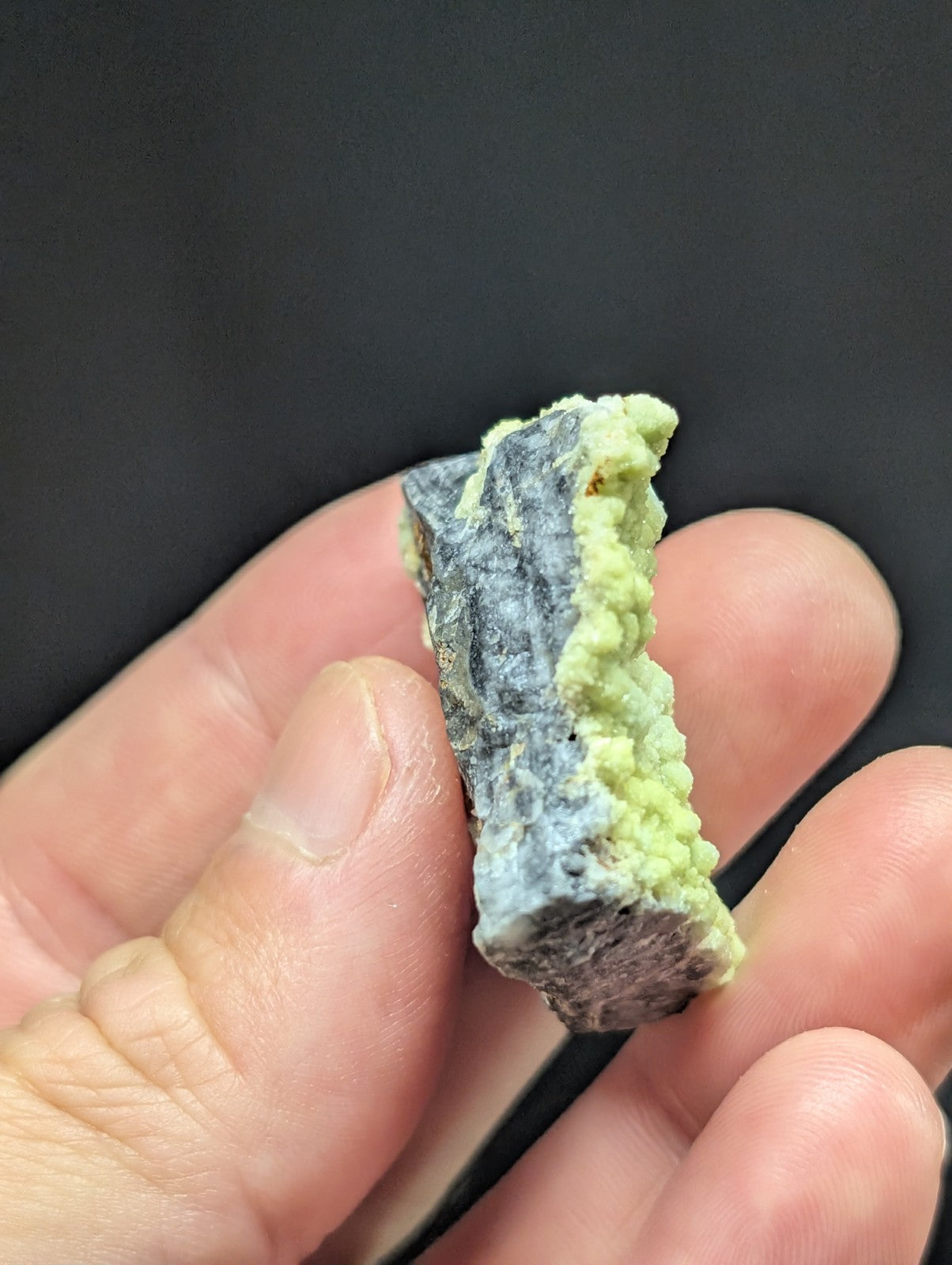A hand holds a small, irregularly shaped mineral specimen featuring a dark gray base with vibrant green crystalline growths along its edge.