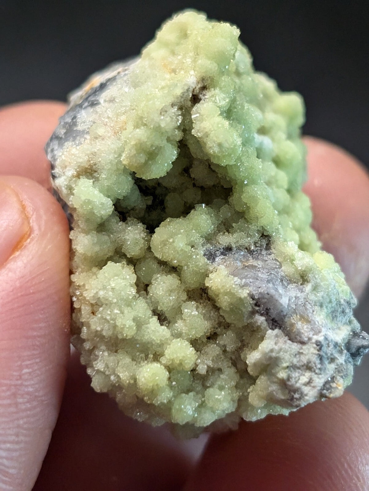 A cluster of pale green, crystalline minerals with a rough, textured surface and some darker grayish matrix.