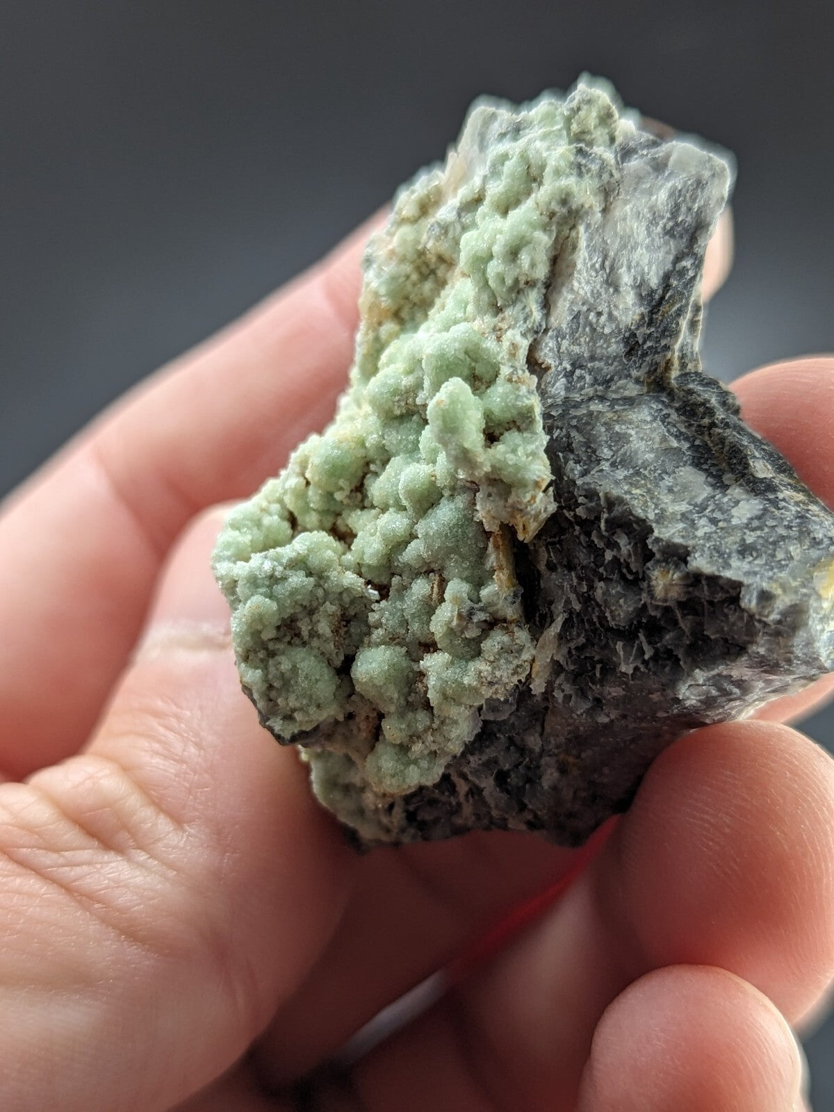 A hand holds a rough, irregularly shaped mineral specimen featuring a vibrant light green crystalline growth against a dark gray matrix.