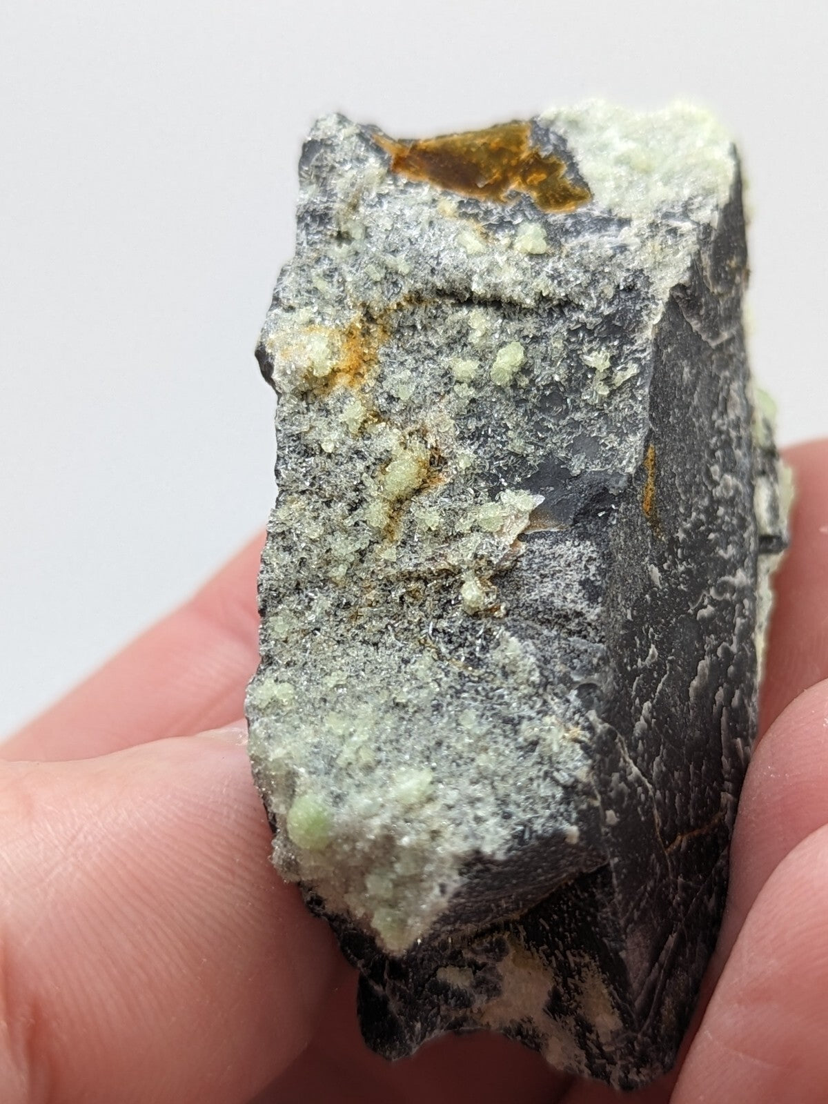 A rough, irregularly shaped mineral specimen with a dark gray base and patches of light green crystals interspersed with small orange-yellow inclusions.
