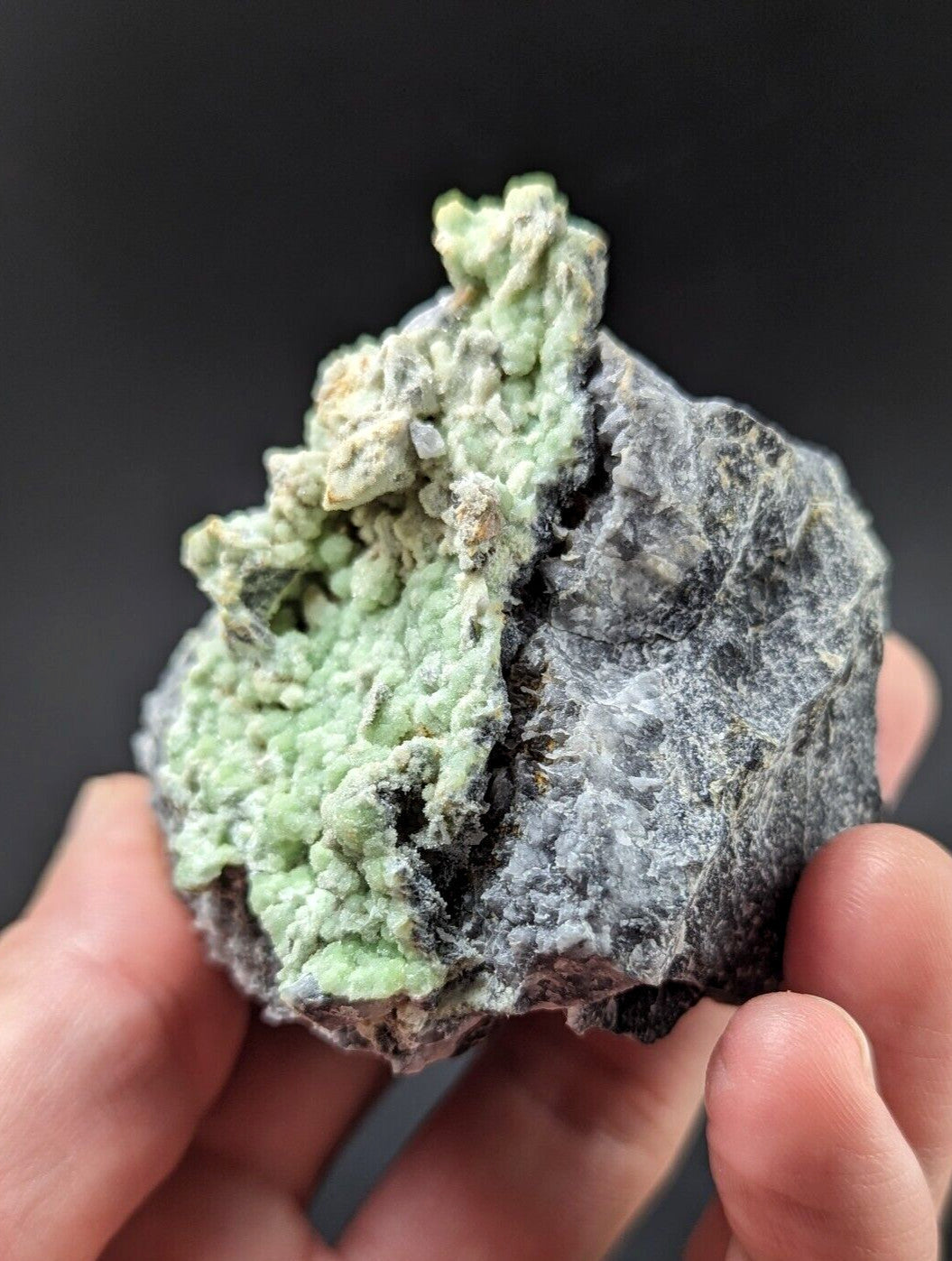 A hand holds a rough, irregularly shaped mineral specimen featuring vibrant light green crystalline formations growing over a dark gray rock base.