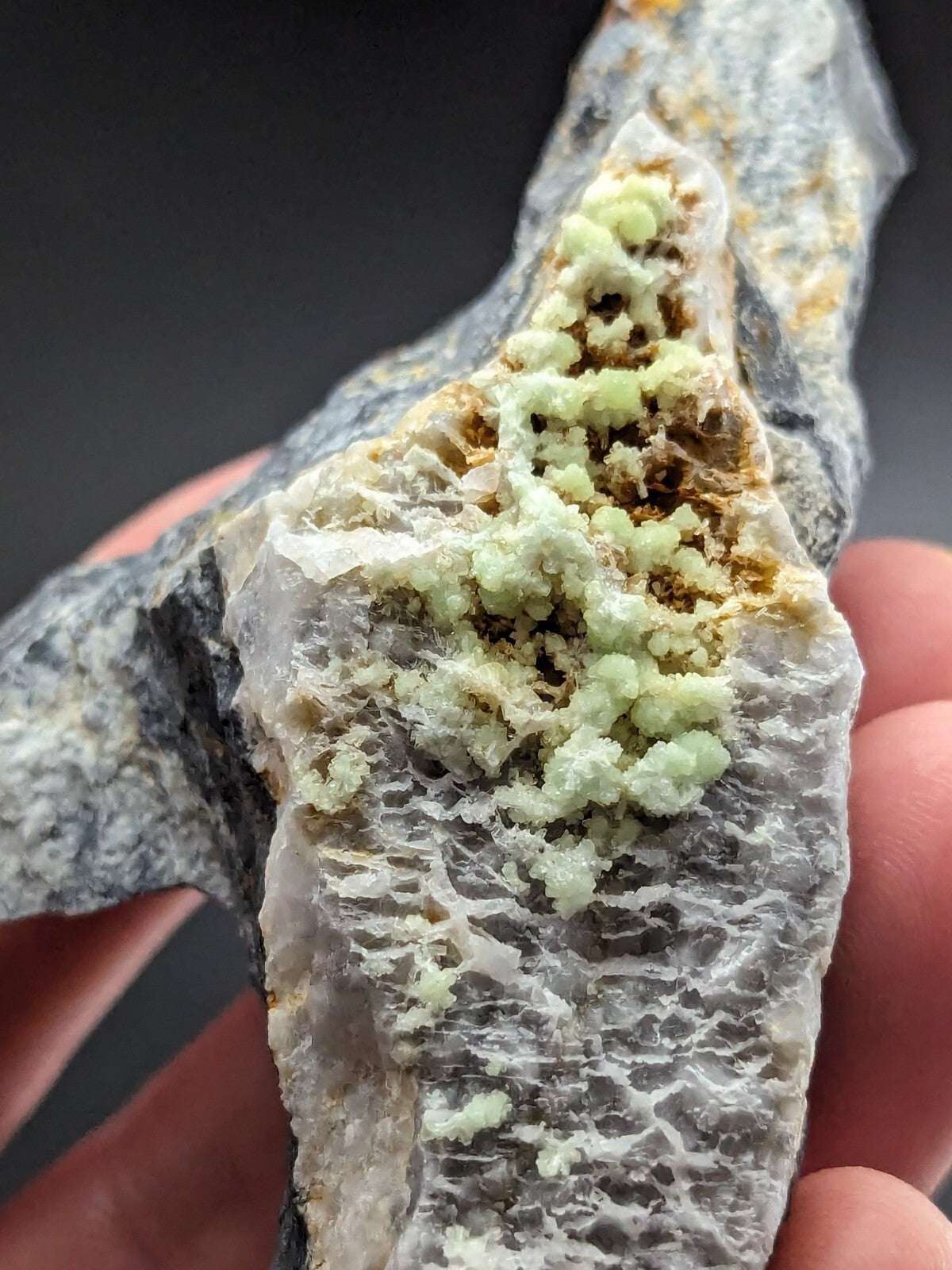 A hand holds a geological specimen featuring clusters of pale green, crystalline mineral formations growing on a grayish rock matrix with visible veining and patches of yellowish material.