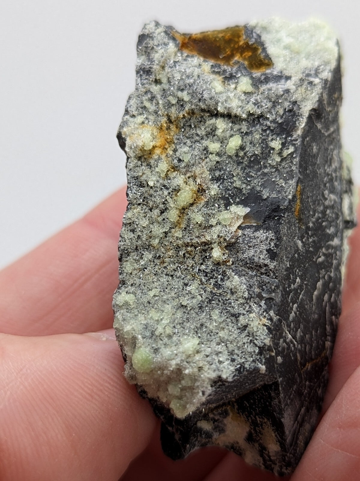 A rugged, irregularly shaped mineral specimen with a dark gray base and speckles of pale green crystals interspersed with patches of golden-yellow inclusions.
