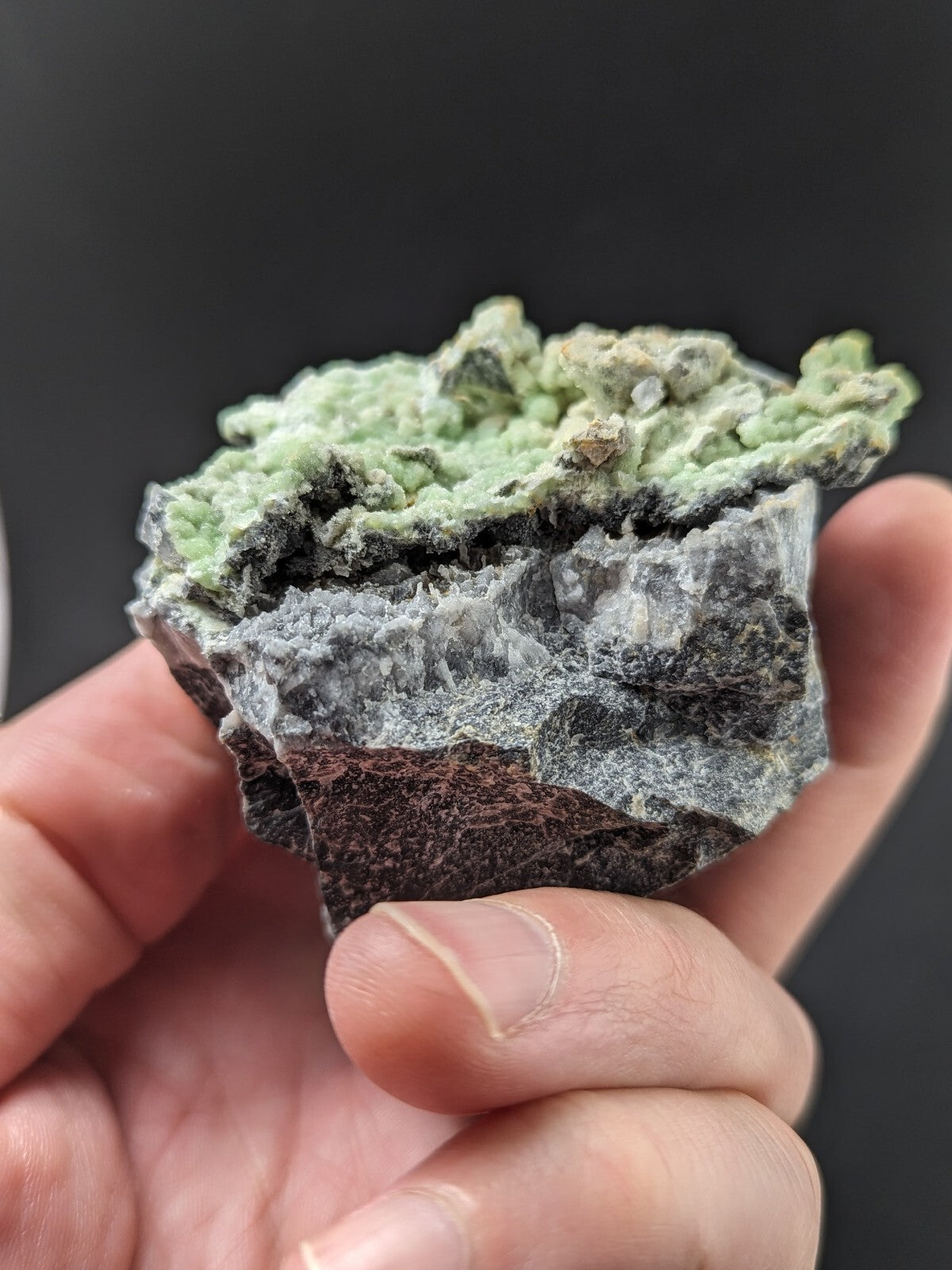 A hand holds a natural mineral specimen featuring vibrant green, textured crystals growing atop a layered base of dark gray and reddish-brown rock.