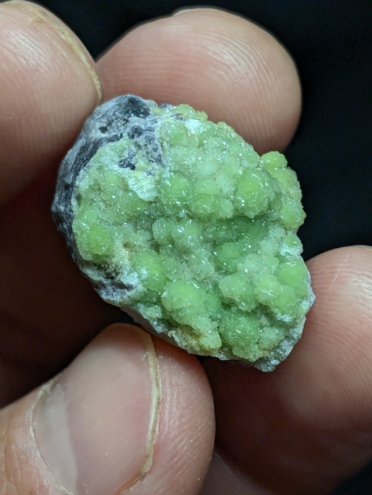 A vibrant green, crystalline mineral specimen with a rough, textured surface and patches of dark gray matrix.