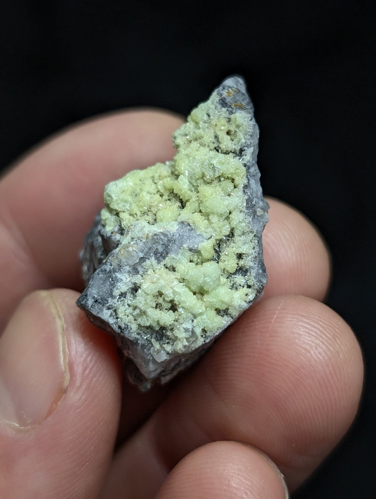 A hand holds a rough, irregularly shaped mineral specimen featuring patches of pale green crystalline growths on a darker gray matrix.