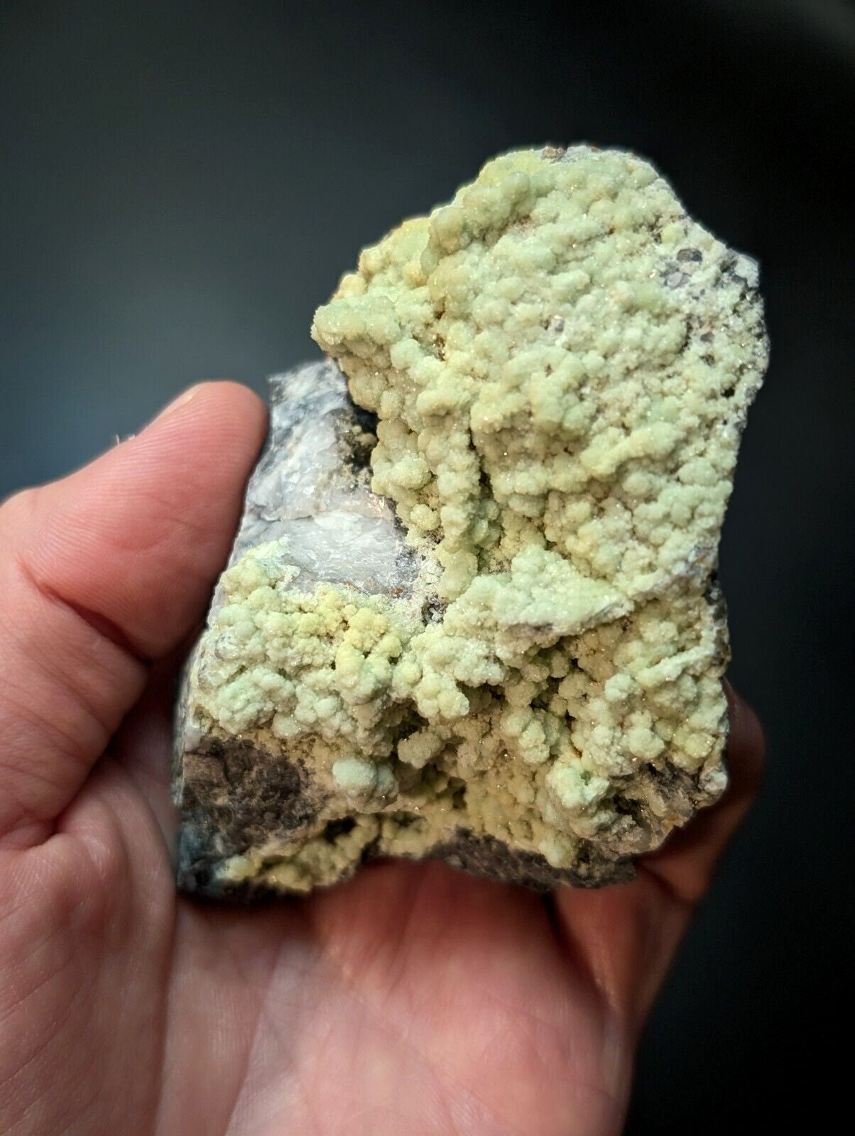 A hand holds a rough, irregularly shaped mineral specimen featuring clusters of pale greenish-yellow crystals growing over a darker gray rock base.