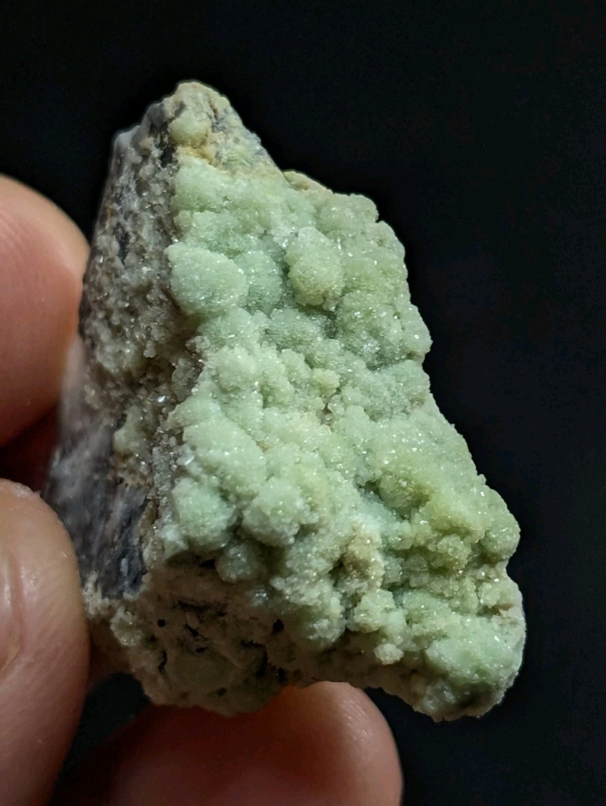 A pale green, crystalline mineral specimen with a rough, textured surface and embedded darker matrix.