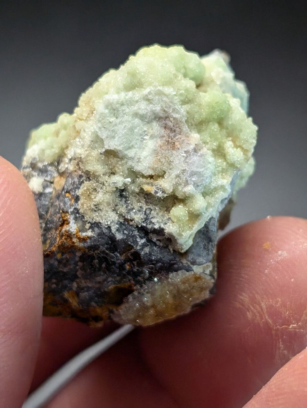A textured, pale green mineral specimen with a rough, crystalline surface and patches of darker gray and yellowish material.