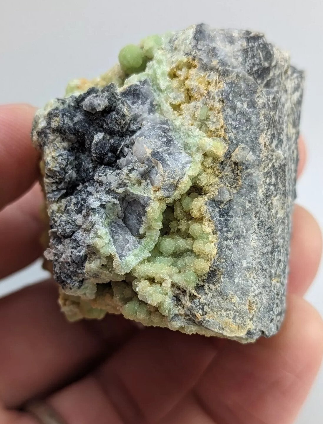 A hand holds a rough, irregularly shaped mineral specimen featuring patches of dark gray rock with vibrant green and yellow crystalline formations.
