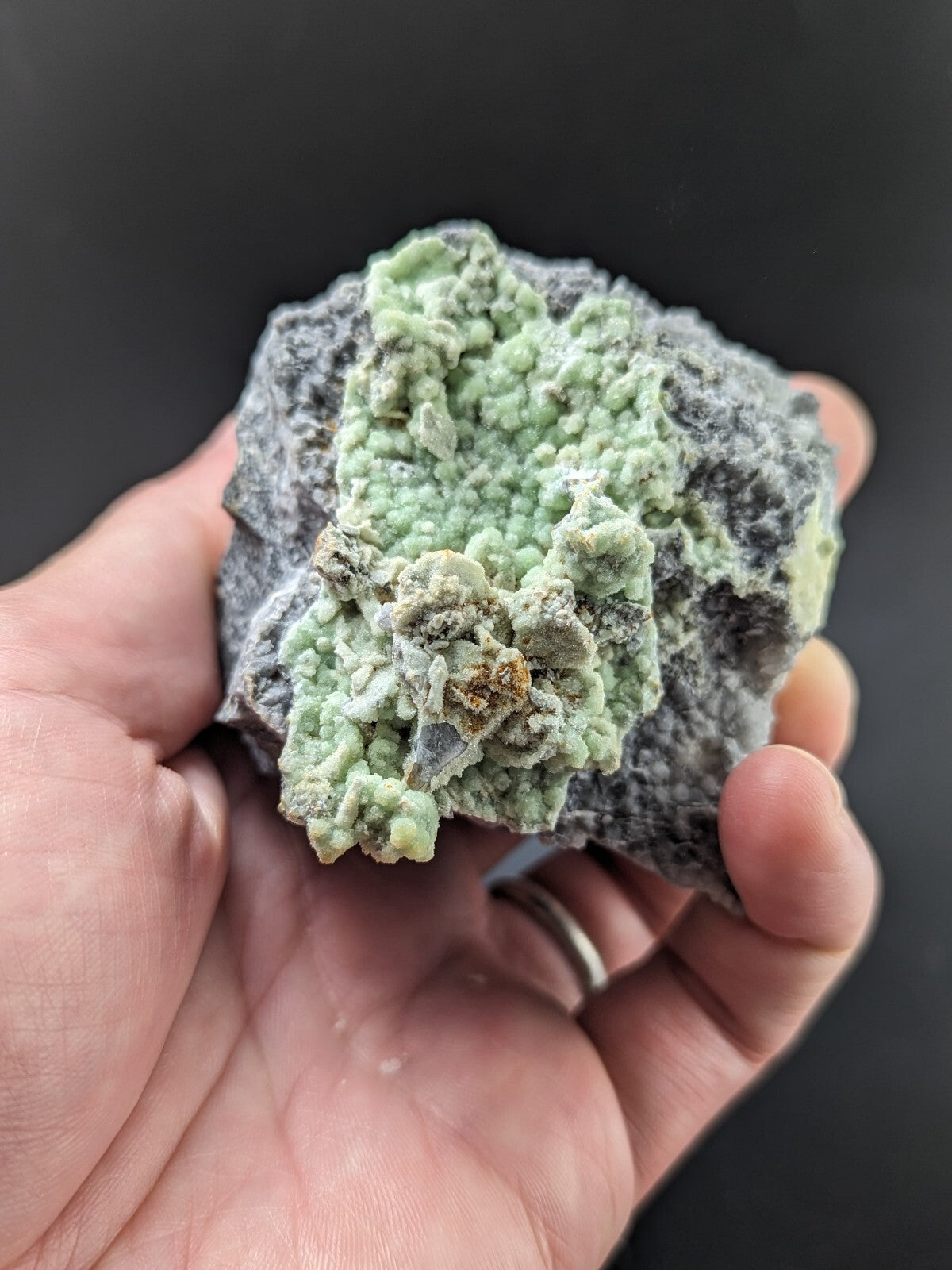 A hand holds a rough, irregularly shaped rock featuring prominent light green mineral formations with textured, bumpy surfaces against a darker gray matrix.