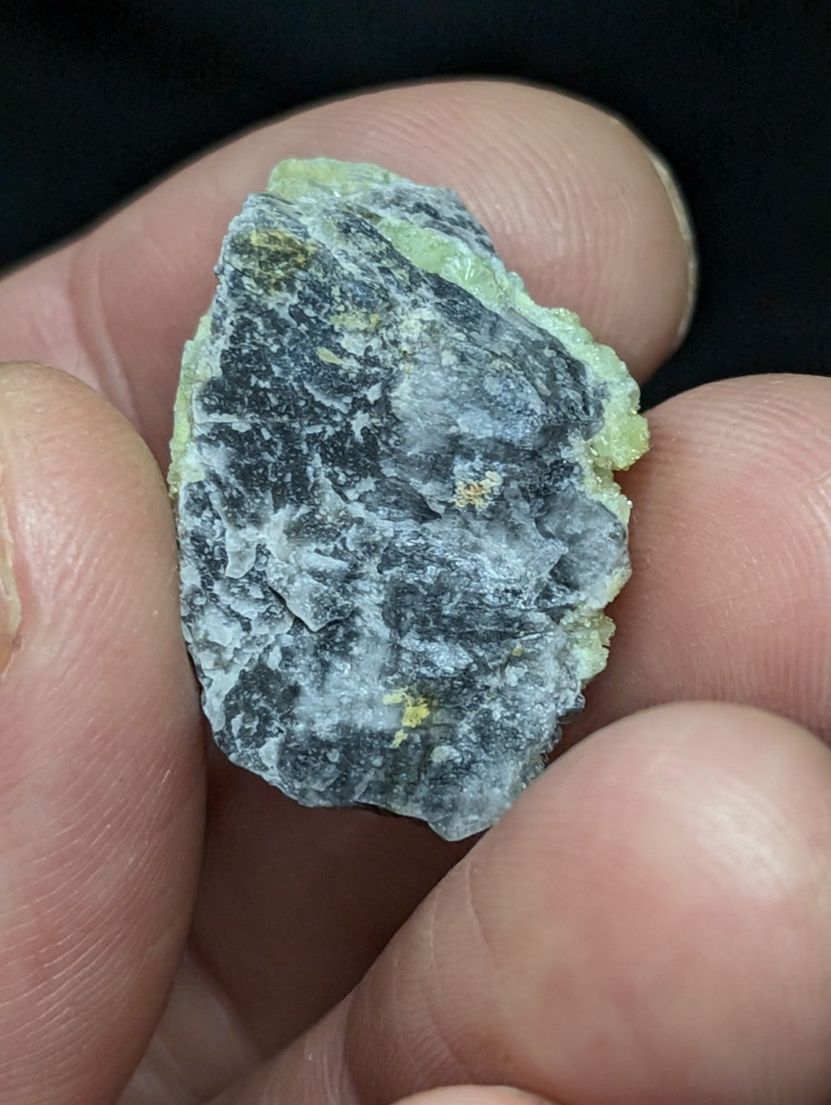 A rough, irregularly shaped mineral specimen with a mottled gray and pale green surface, featuring subtle yellowish specks.