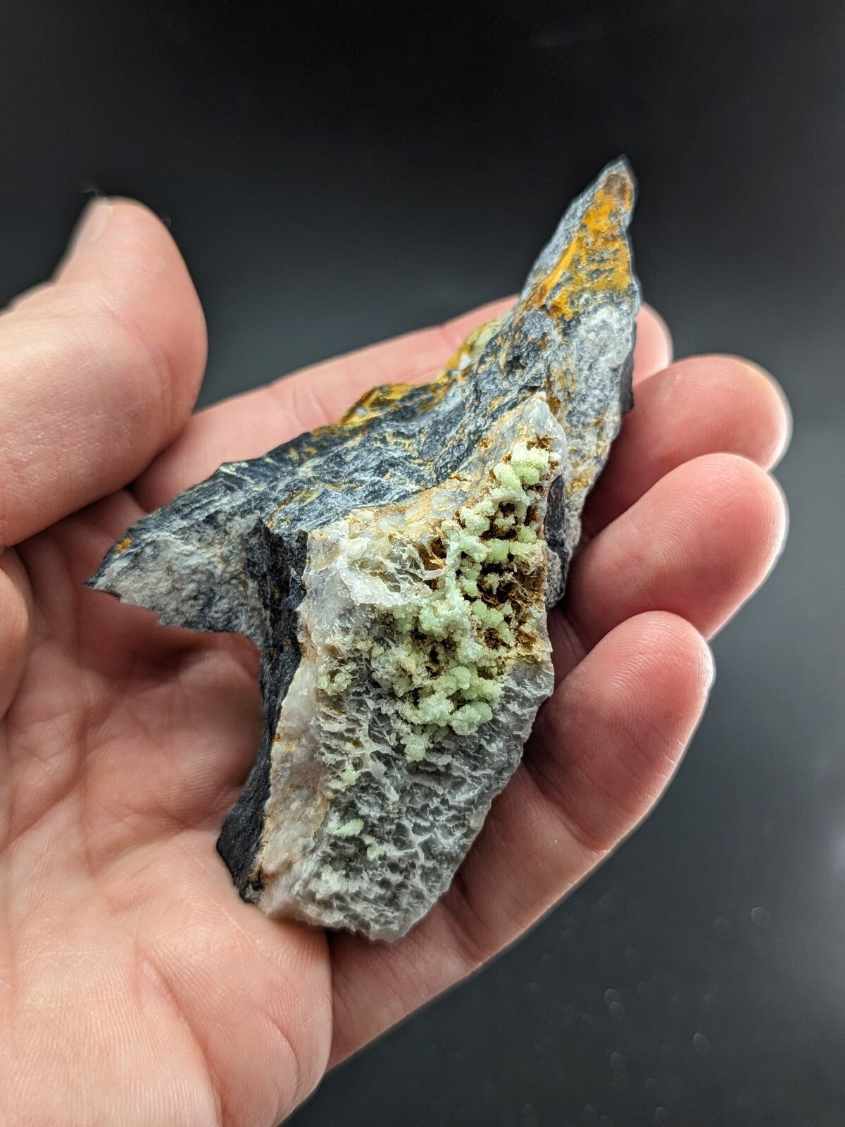 A hand holds a rugged, dark gray rock specimen featuring patches of vibrant green and yellow mineral growths.