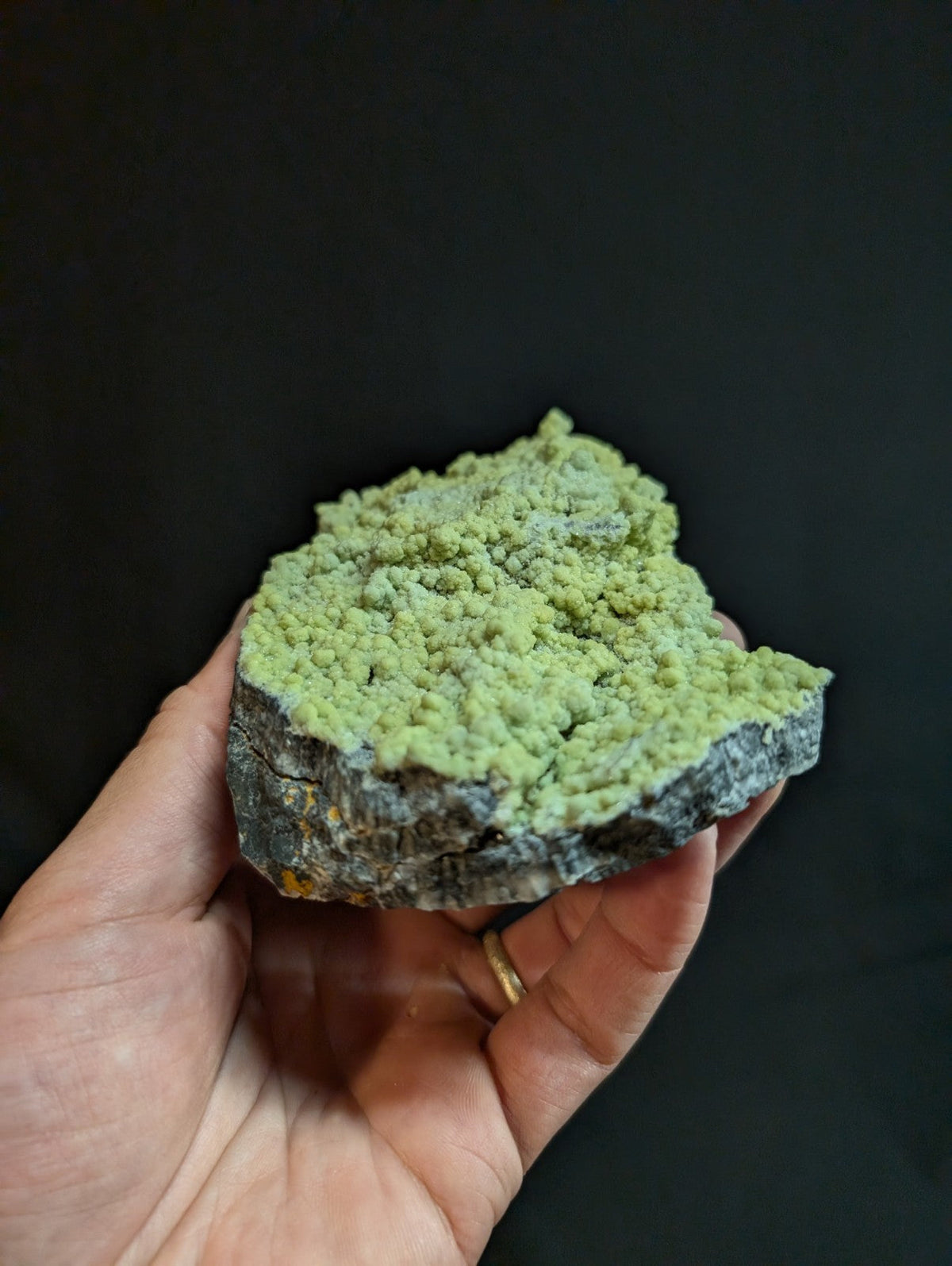 A hand holds a rough, irregularly shaped mineral specimen featuring a vibrant green crystalline growth atop a darker, textured base.