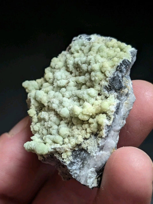 A hand holds a rough, irregularly shaped mineral specimen featuring clusters of pale greenish-yellow crystals growing from a darker gray and white rock matrix.