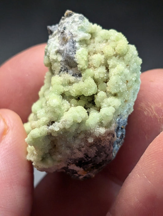 A pale green, crystalline mineral specimen with a rough, textured surface and patches of darker matrix.